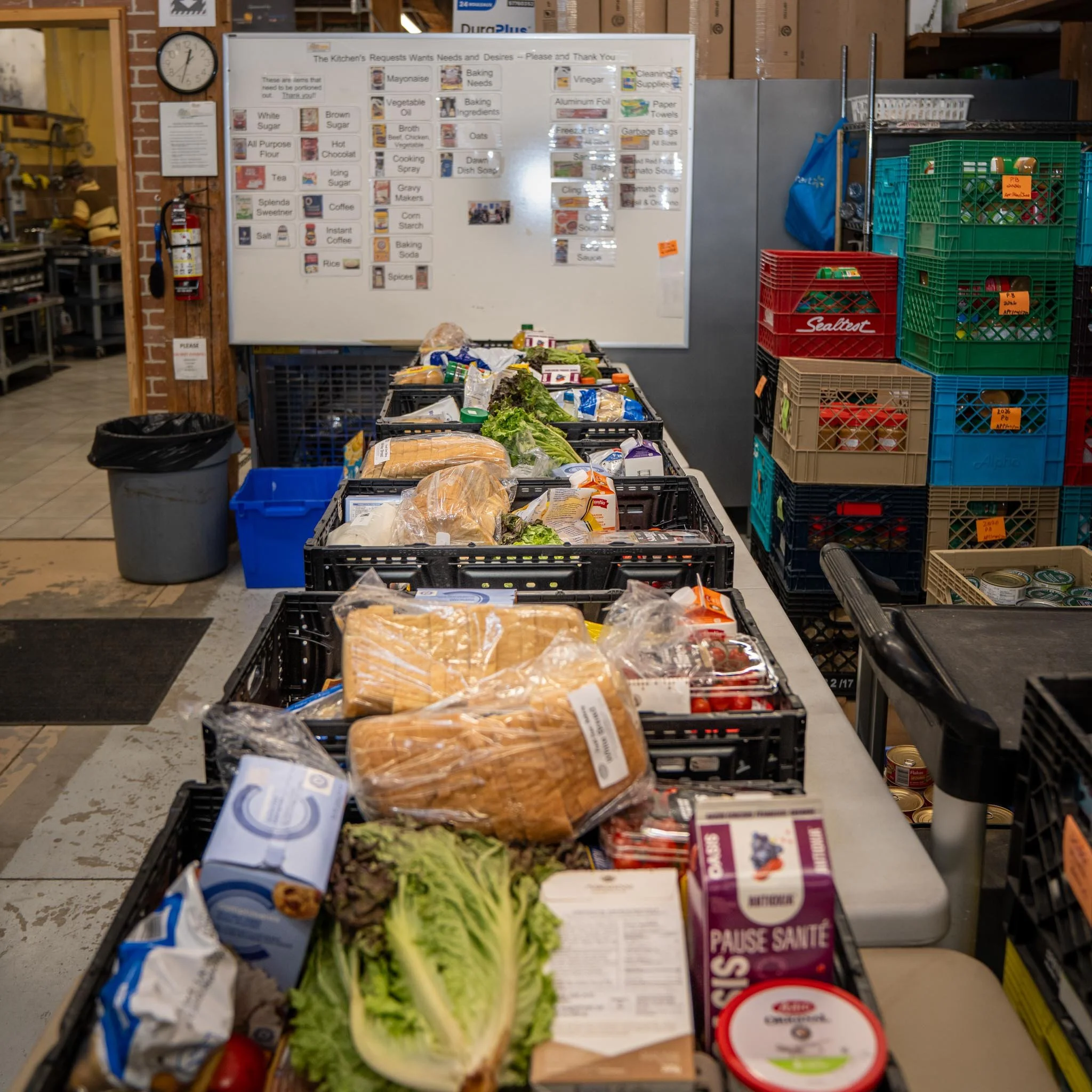 A grocery store checkout counter with various food items in black plastic crates including bread, lettuce, and packaged products. In the background, there are colorful plastic crates stacked on each other and a whiteboard with a list of kitchen needs and desires.