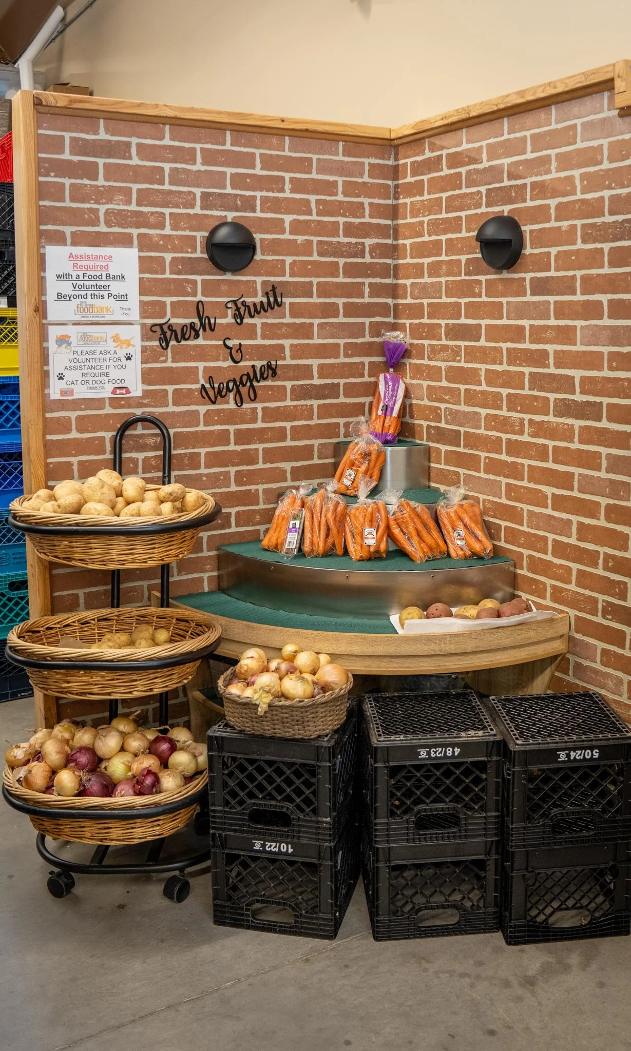 Display of fresh onions, carrots, and potatoes at a food bank with a brick wall background and signs indicating assistance for food donations.