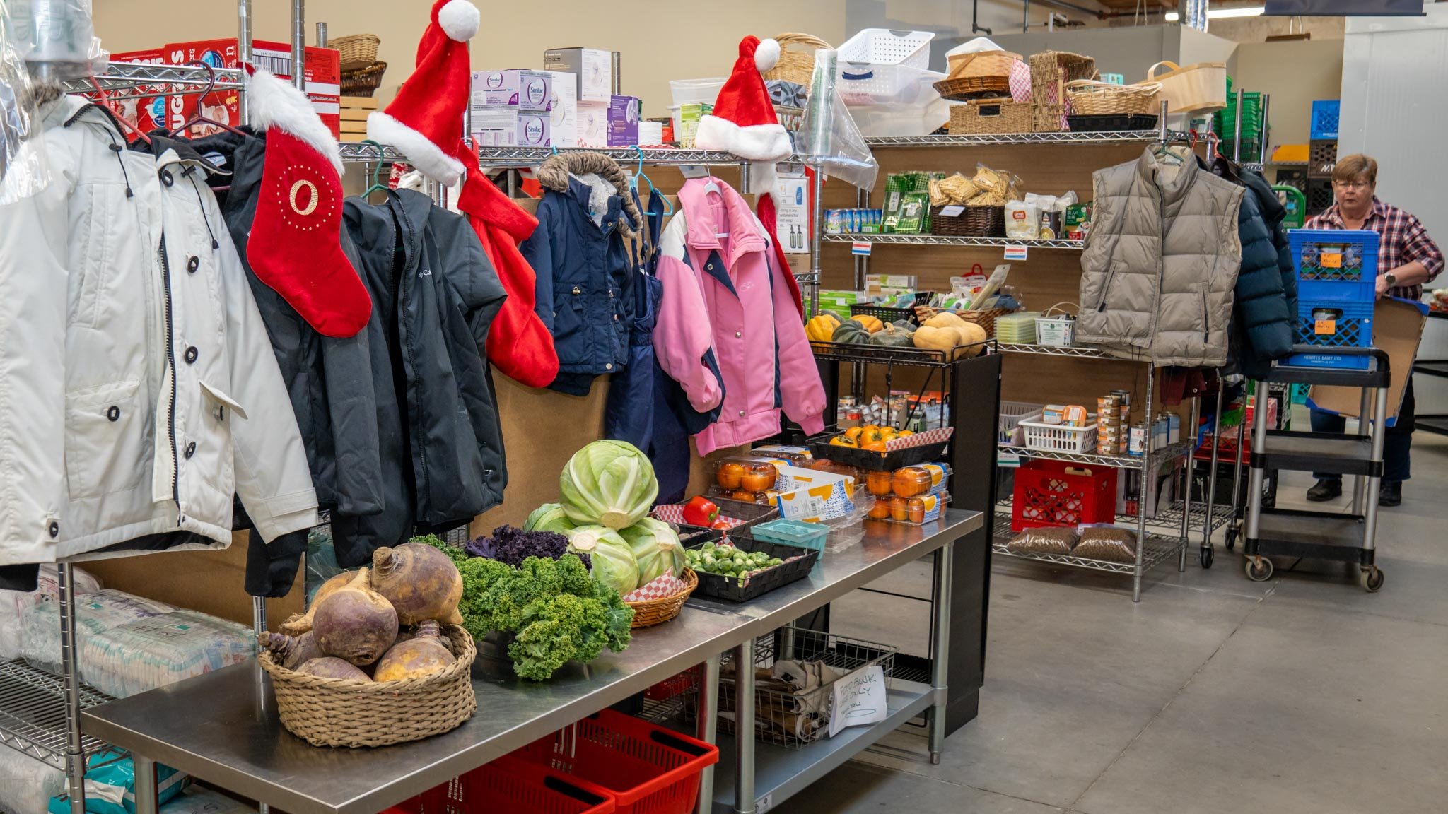 A thrift store or charity shop display with winter jackets, Santa hats, and seasonal decorations, alongside fresh vegetables and produce on metal tables and shelves.