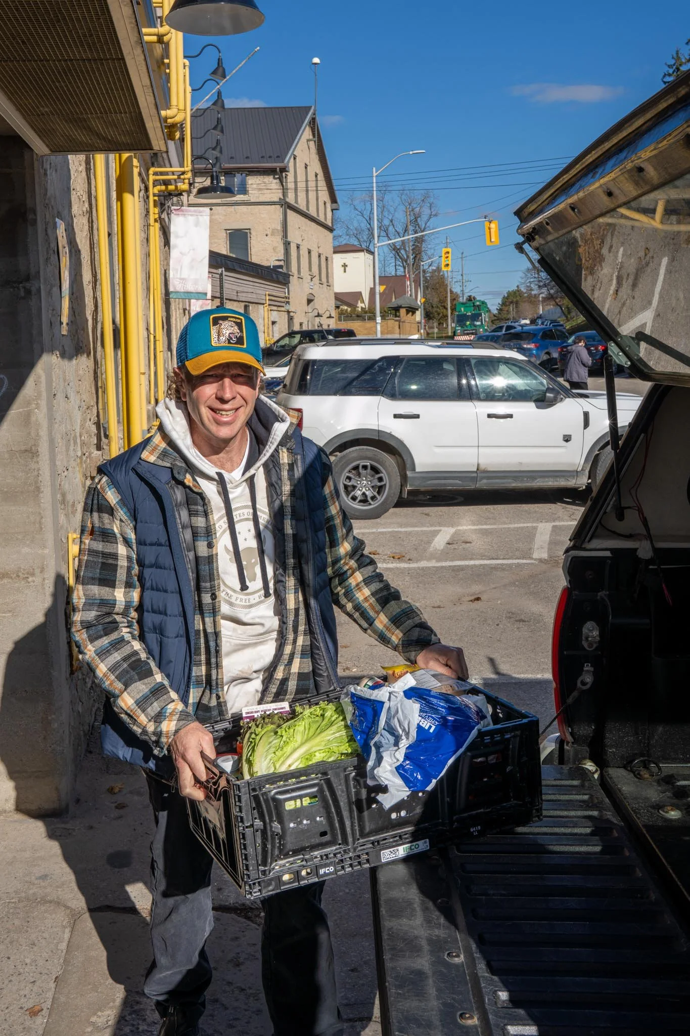 A man standing outside with a crate of groceries, smiling at the camera. He is wearing a blue baseball cap, a plaid jacket, and a hoodie. The open trunk of a vehicle is visible beside him, and the background shows a parking lot with several cars and a street with traffic lights.