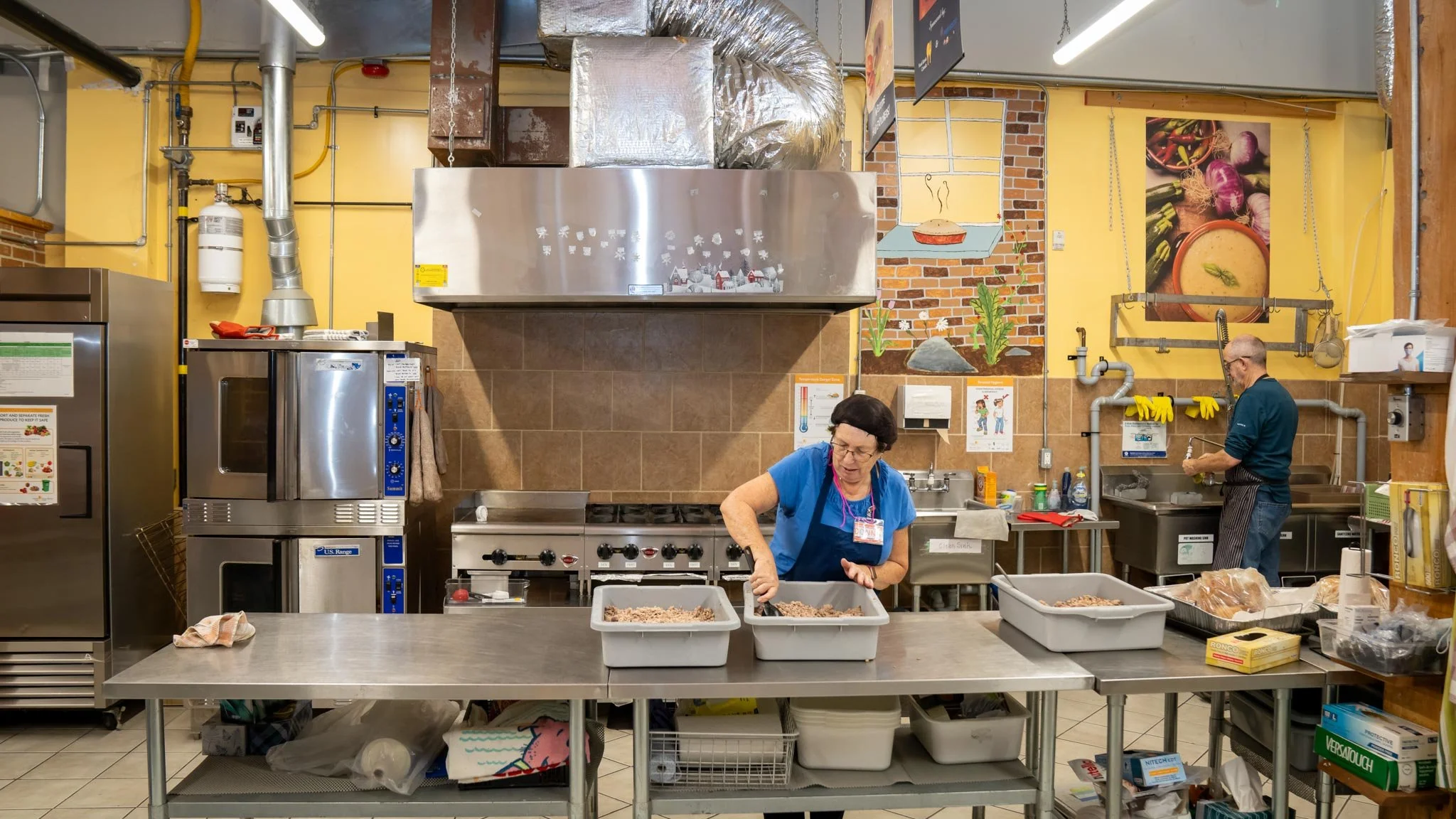 Two people working in a commercial kitchen, preparing food at a stainless steel counter. The woman in the foreground is mixing ingredients in two plastic containers, and the man in the background is washing dishes at a sink. The kitchen has yellow walls, brick accents, and various kitchen appliances and supplies.