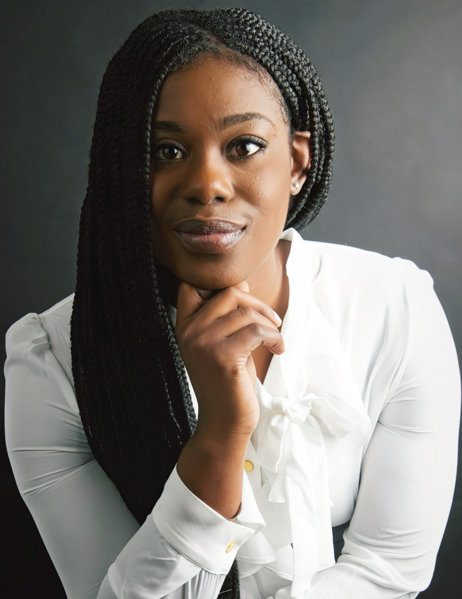 A woman with dark braided hair, brown eyes, and natural makeup, wearing a white outfit, resting her chin on her hand, looking at the camera, against a dark background.