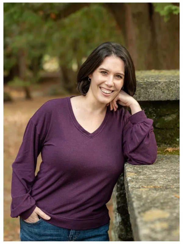 A woman with shoulder-length dark hair, wearing a purple long-sleeve shirt, smiling and leaning on a stone ledge outdoors in a park setting with trees in the background.
