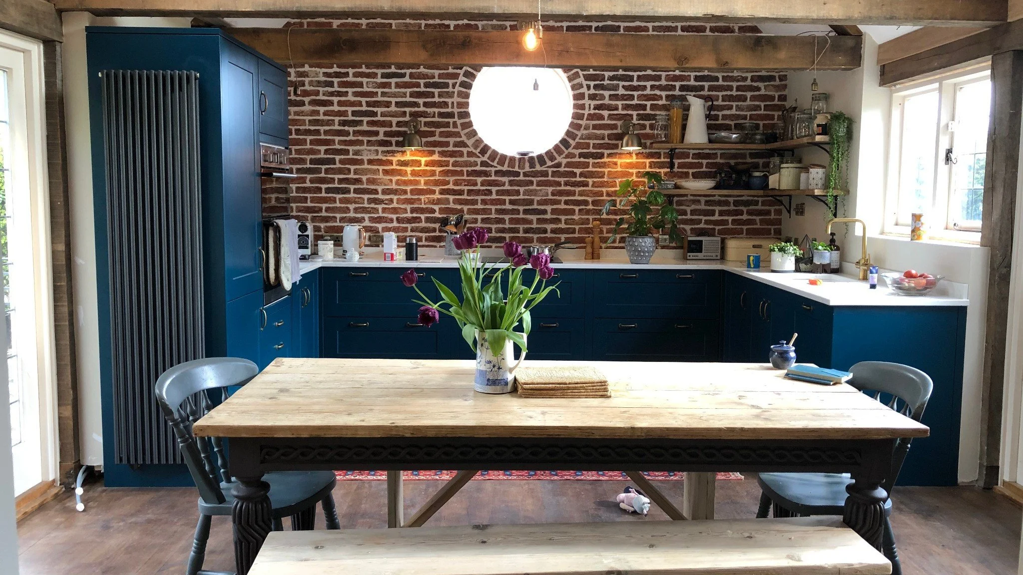 Modern rustic kitchen-diner featuring exposed timber beams, red brick wall, blue shaker cabinets, and a wooden farmhouse table with flowers.