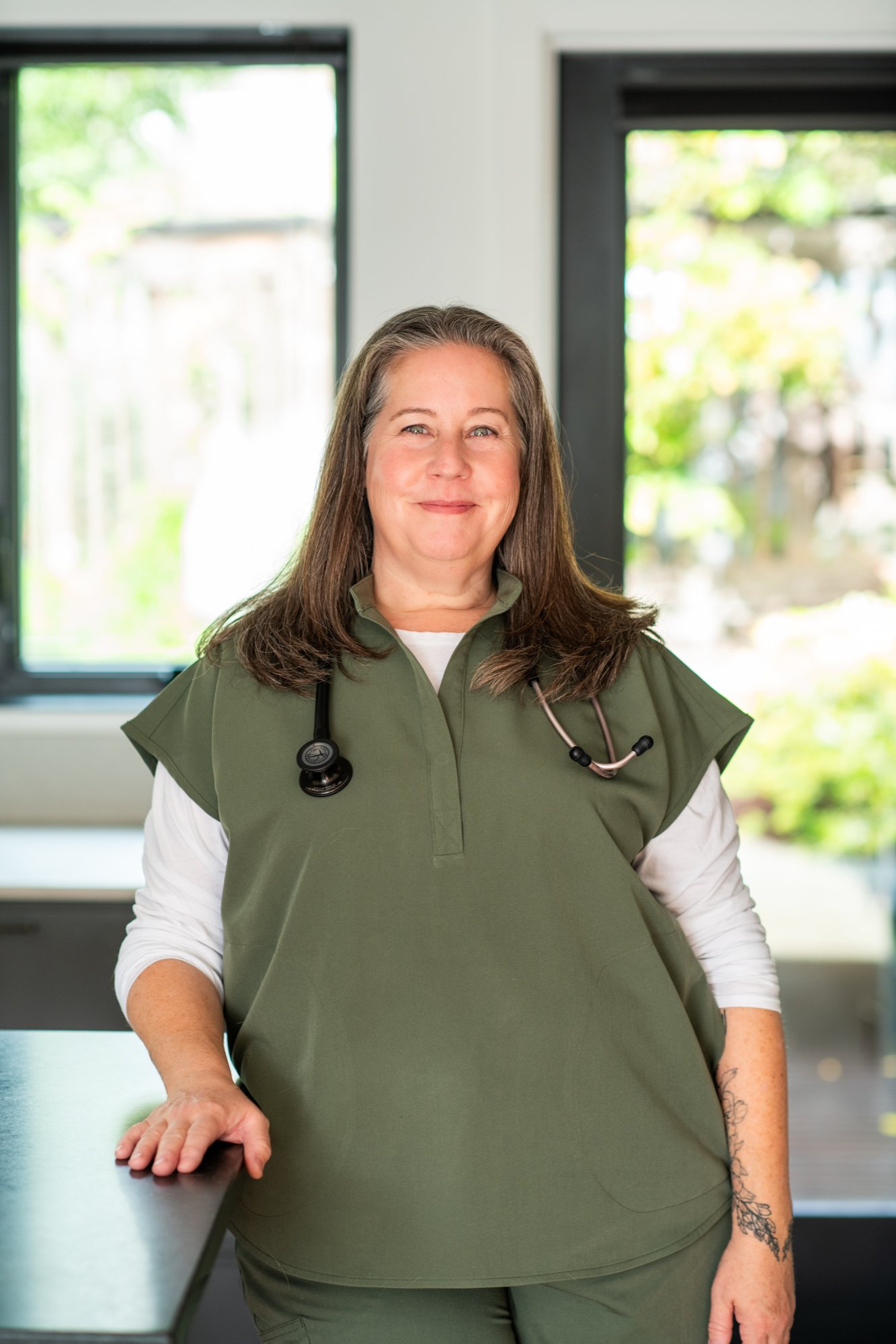 Smiling female healthcare worker in green scrubs with stethoscope around neck, standing in bright medical office.