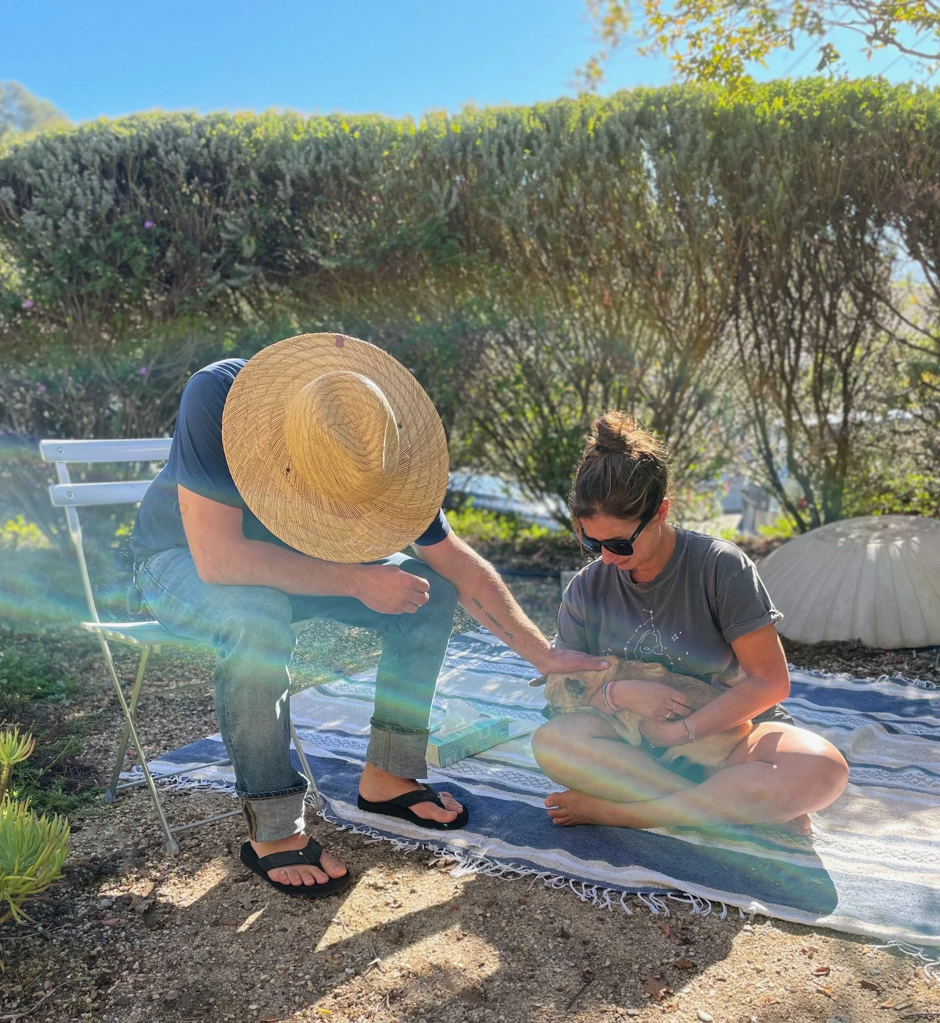 A woman sitting on a blanket with a dog in her lap, and a man wearing a large straw hat petting the dog outdoors on a sunny day with bushes and trees in the background.