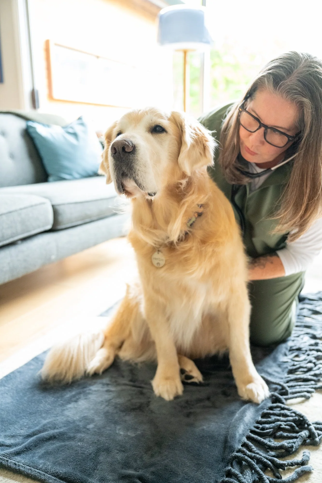 A woman with glasses and long hair kneels next to a sitting golden retriever dog on a black rug in a living room, with a gray sofa and cushions in the background.