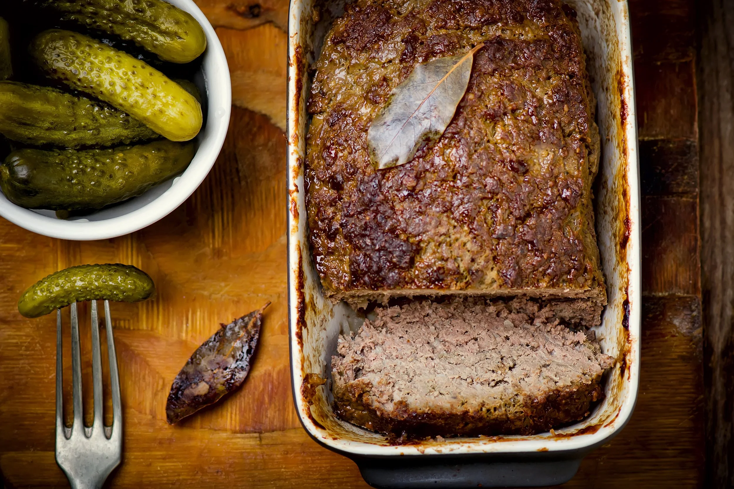Terrine de pâté de viande cuite au four dans un plat rectangulaire, coupée en tranches, décorée d'une feuille de laurier. Cornichons marinés et fourchette vintage.