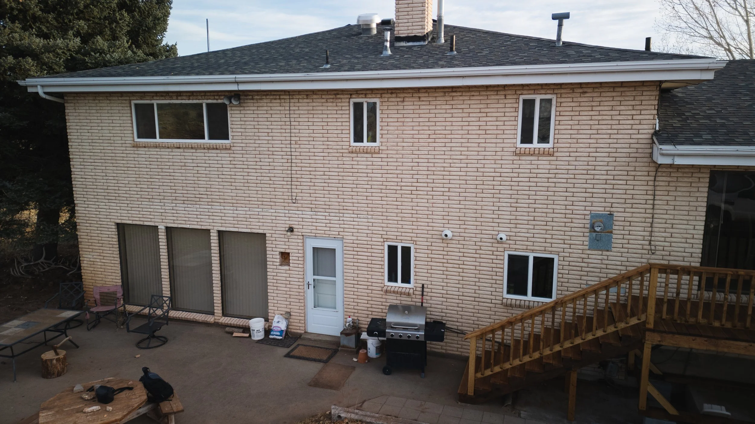 Rear view of a two-story beige brick house with several windows, a small deck with wooden stairs, outdoor grill, and patio furniture. Real estate drone photography and videography services in Roosevelt, Vernal, Uintah Basin, Utah, and nationwide.