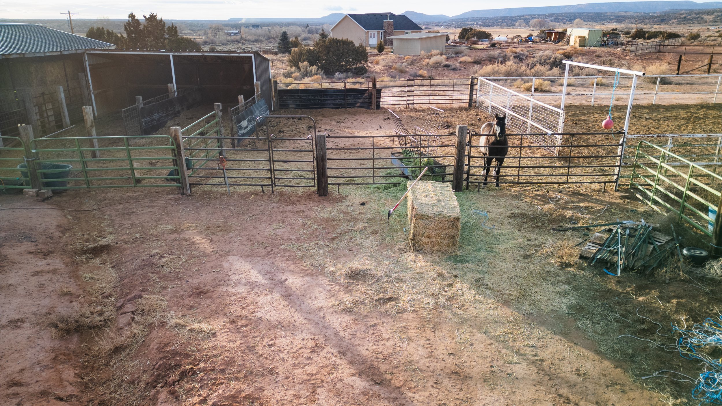 A rural farm scene with multiple fenced areas, a single horse standing near the fence, hay bales, and farming equipment. Real estate drone photography and videography services in Roosevelt, Vernal, Uintah Basin, Utah, and nationwide.