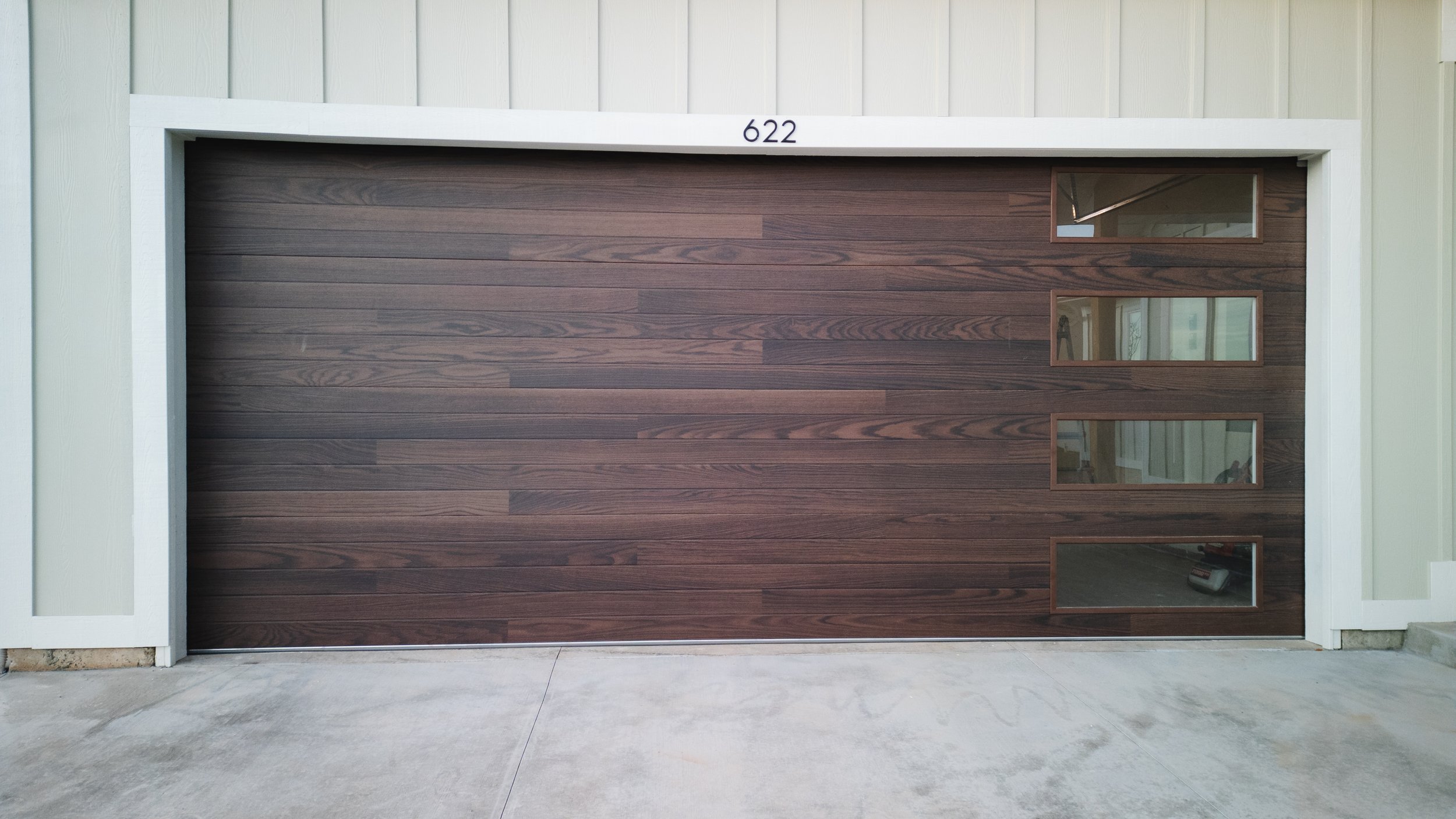 Modern brown wooden garage door with four rectangular windows on the right side, framed with brown wood, in a white exterior wall. Real estate drone photography and videography services in Roosevelt, Vernal, Uintah Basin, Utah, and nationwide.