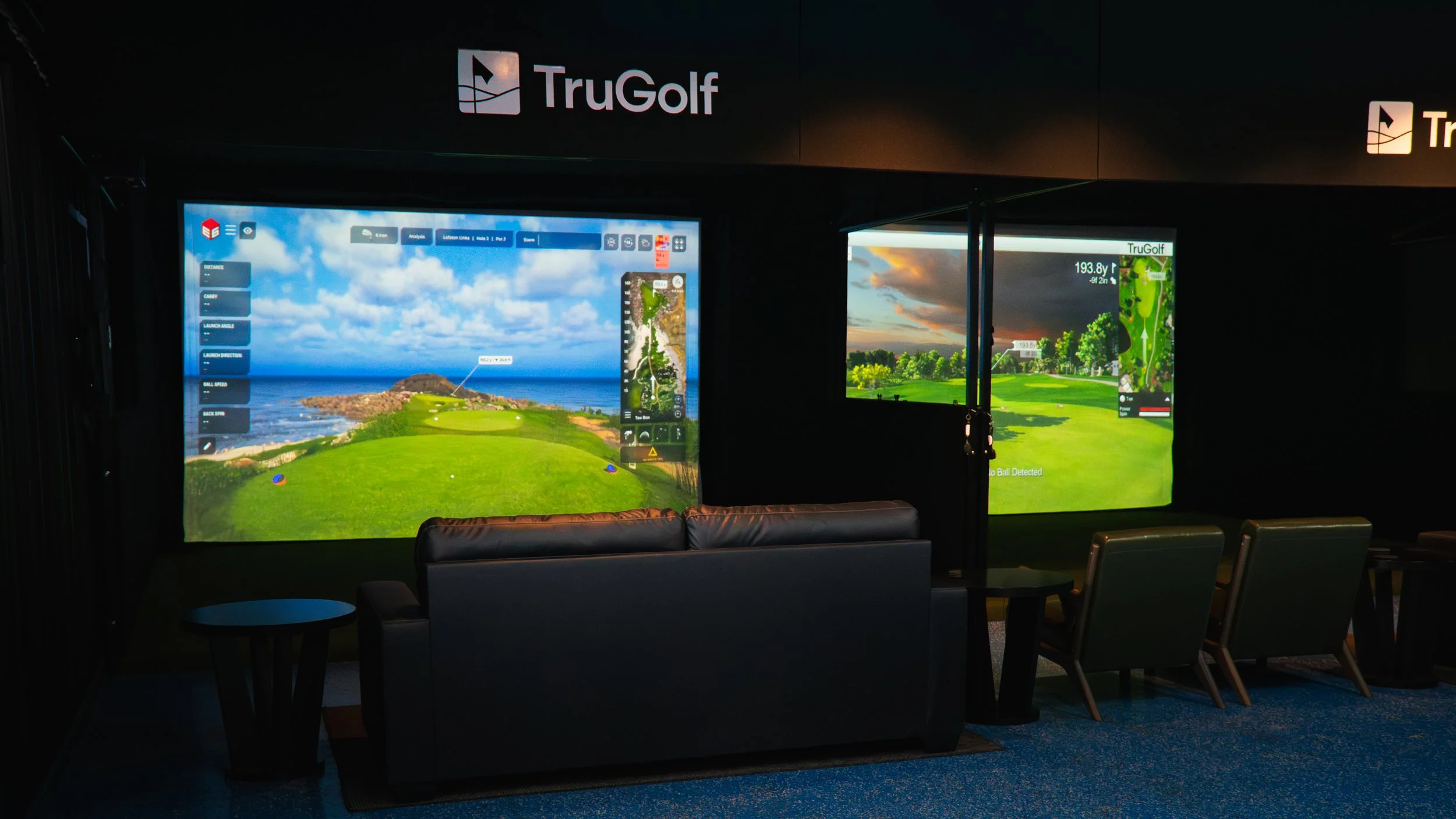 Ground photograph of an indoor golf simulator room with two large screens displaying virtual golf courses, a black sofa, small tables, and green chairs.