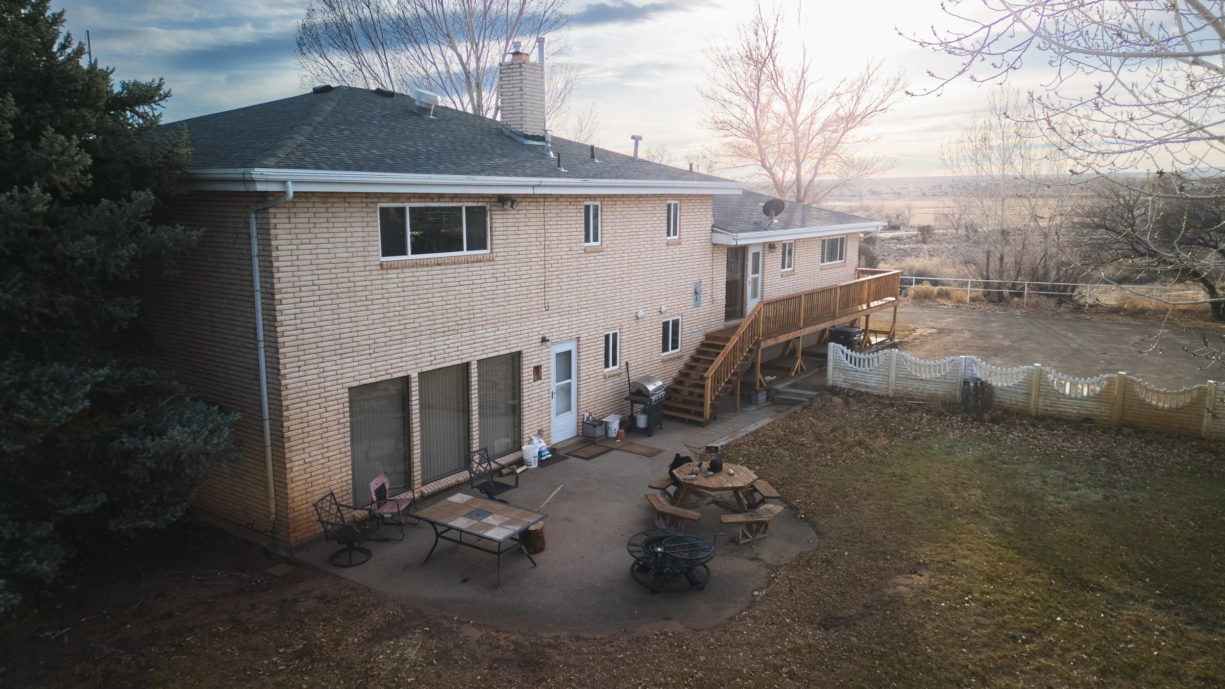 View of the backyard and rear of a two-story brick house with a wooden deck and outdoor seating area, surrounded by trees and open landscape, during sunset. Real estate drone photography and videography services in Roosevelt, Vernal, Uintah Basin, UT