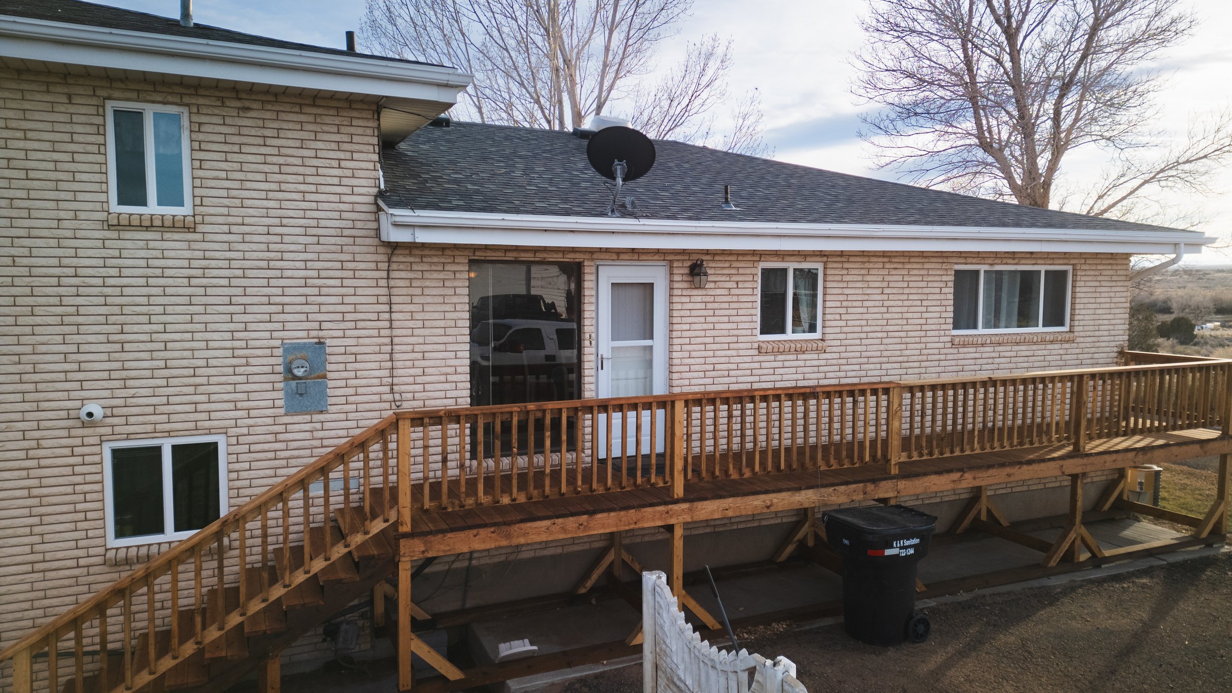 Back of a house with an elevated wooden deck, white brick exterior, multiple windows, a satellite dish, and a white door. There is a black trash bin and some construction materials underneath the deck.