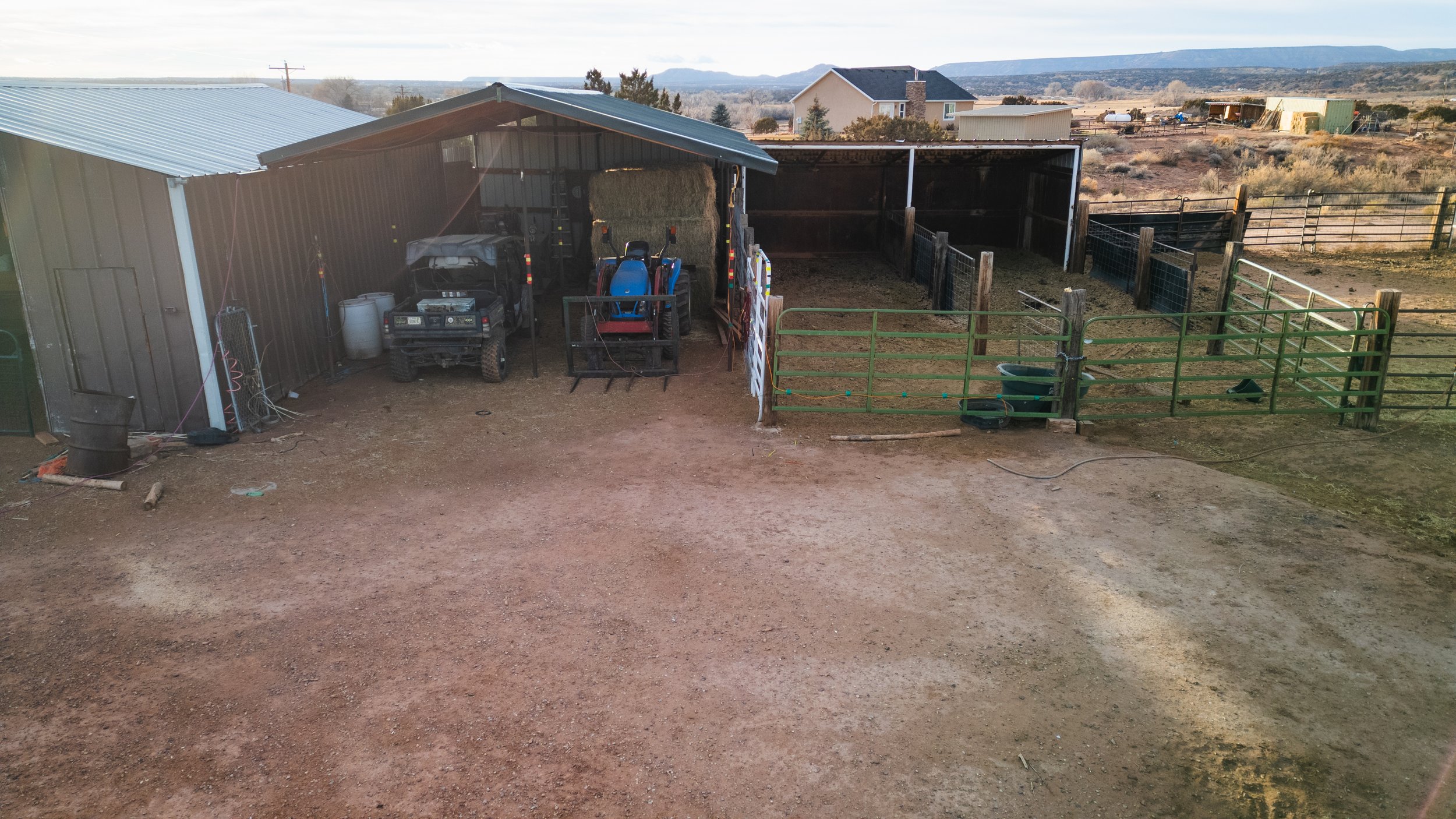 A dirt farm yard with a metal shed containing farming equipment and hay bales, surrounded by fences and open land with houses and mountains in the background. Real estate drone photography and videography services in Roosevelt, Vernal, Uintah Basin, 