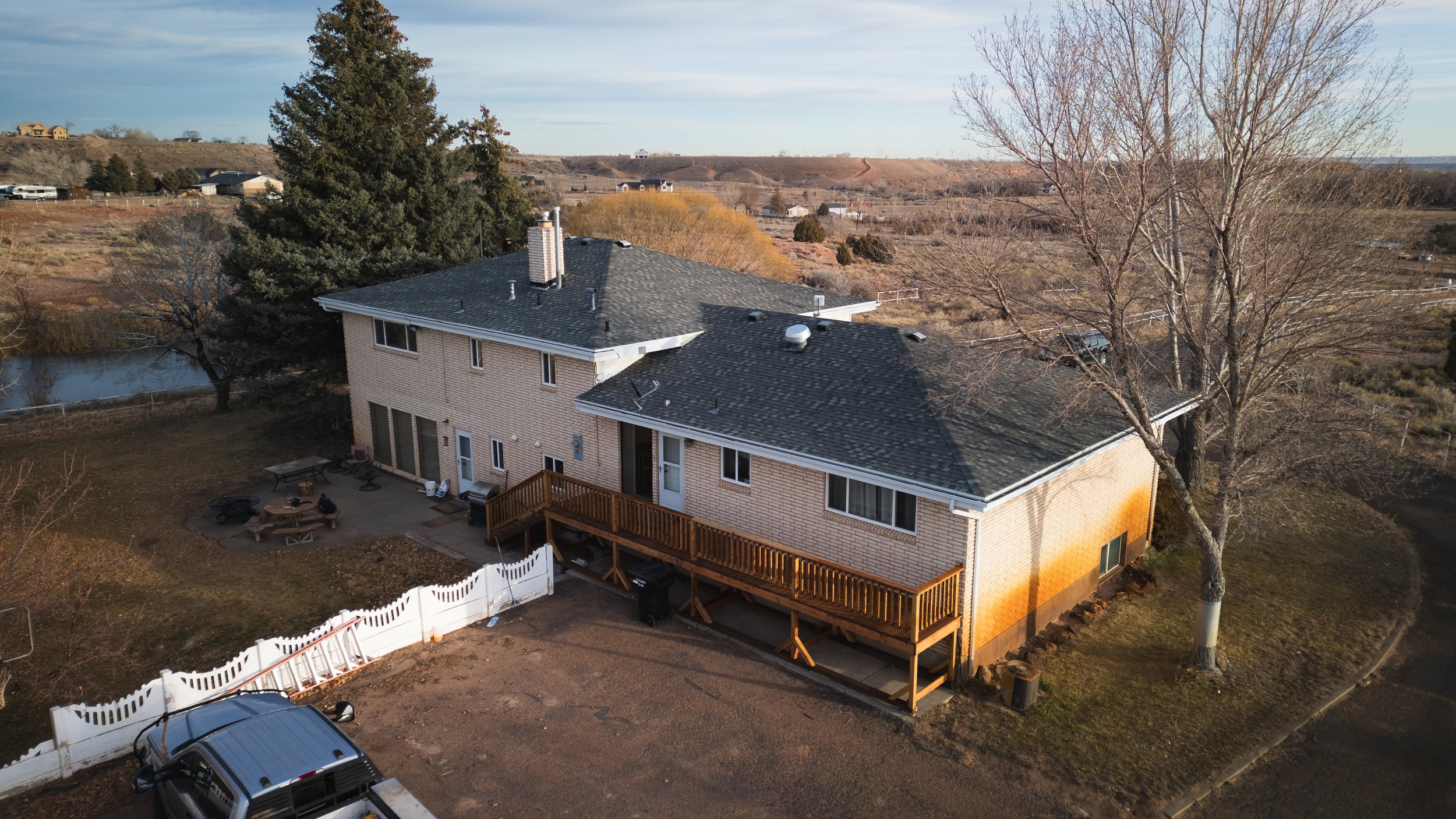 A two-story house with a brick exterior, a wooden deck, and a backyard patio with outdoor furniture, surrounded by leafless trees and a fenced yard. Real estate drone photography and videography services in Roosevelt, Vernal, Uintah Basin, Utah.