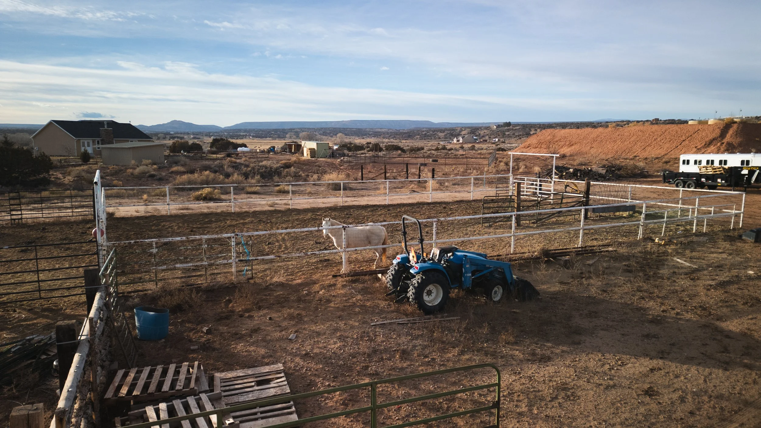 A rural farm scene with a white horse near a fenced area, a blue tractor working the dirt, and some farm buildings in the background. Real estate drone photography and videography services in Roosevelt, Vernal, Uintah Basin, Utah, and nationwide.