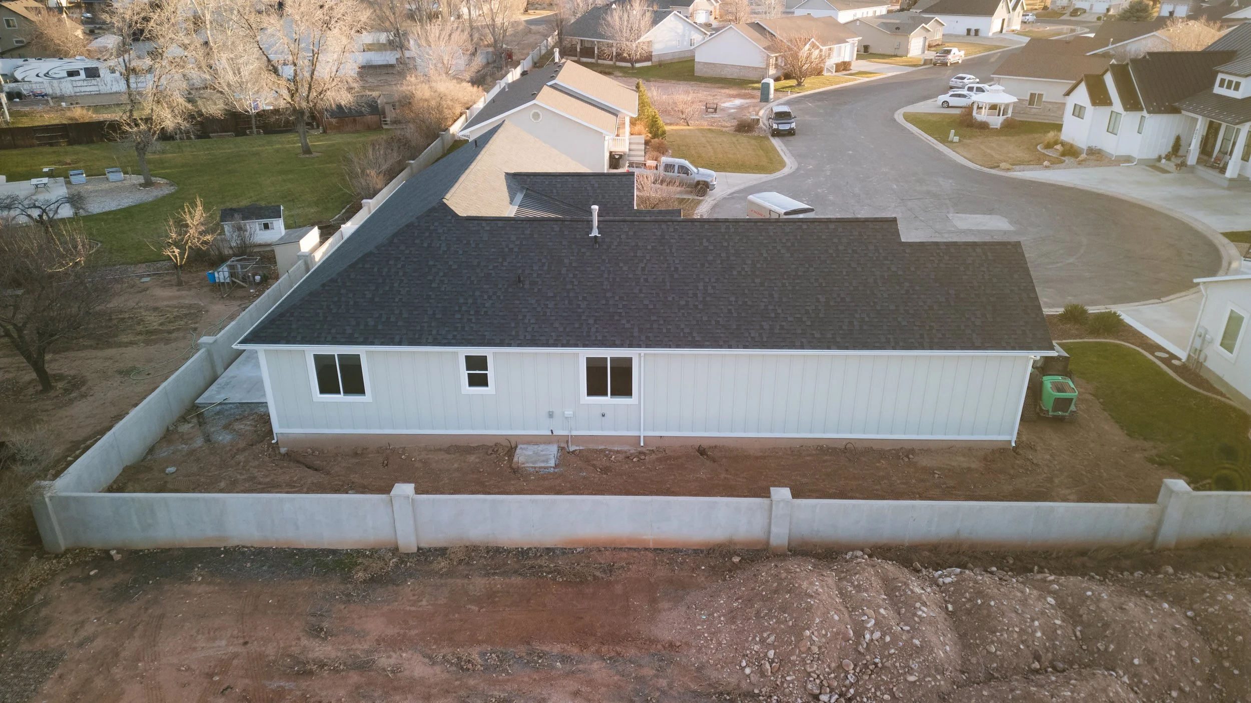 An aerial view of a newly built house with a white exterior, black roof, and fenced backyard in a rural neighborhood. Real estate drone photography and videography services in Roosevelt, Vernal, Uintah Basin, Utah, and nationwide.