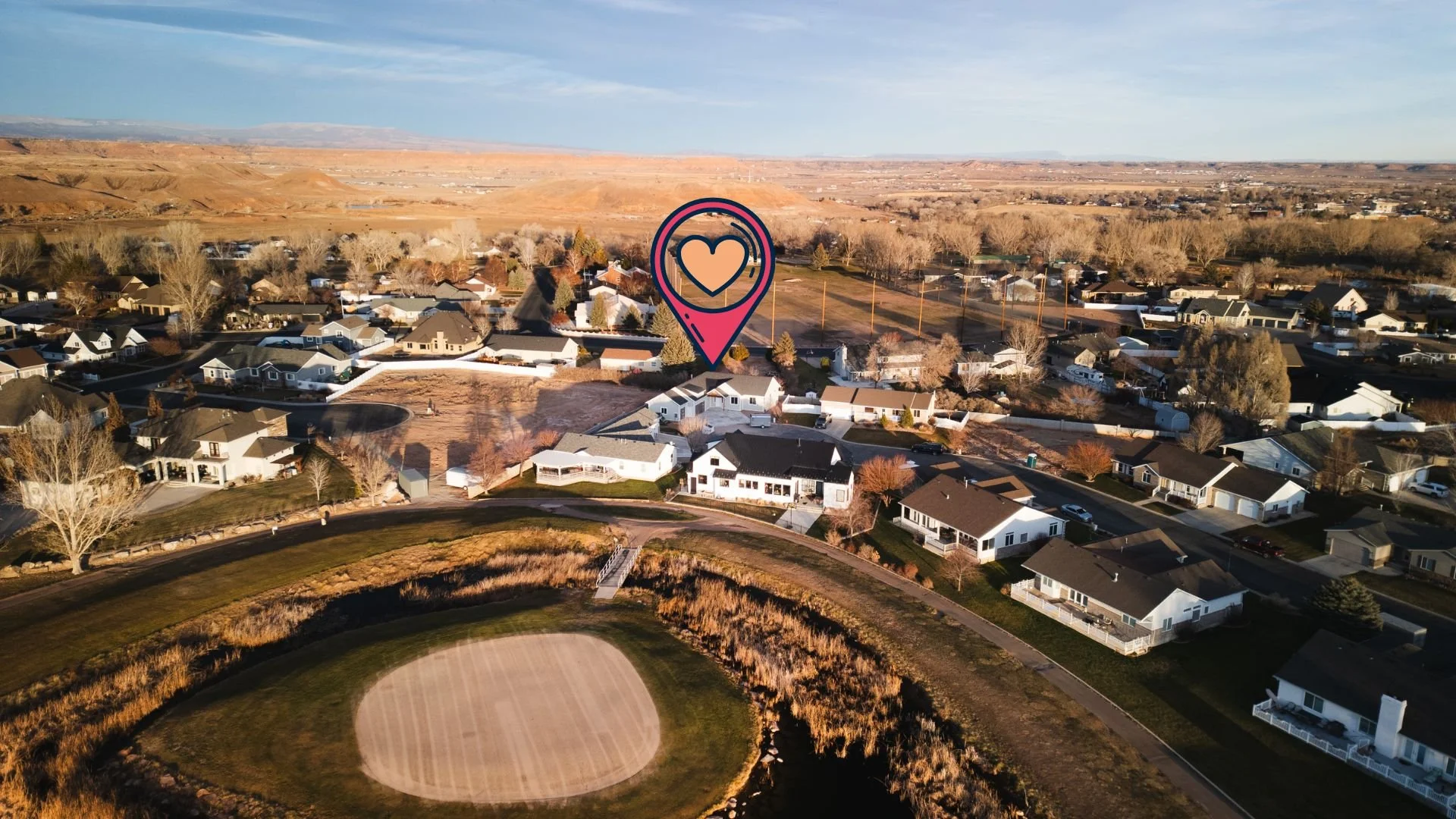Aerial view of a suburban neighborhood with houses, a baseball field, and a marker with a heart icon and location pin over one house. Real estate drone photography and videography services in Roosevelt, Vernal, Uintah Basin, Utah, and nationwide.