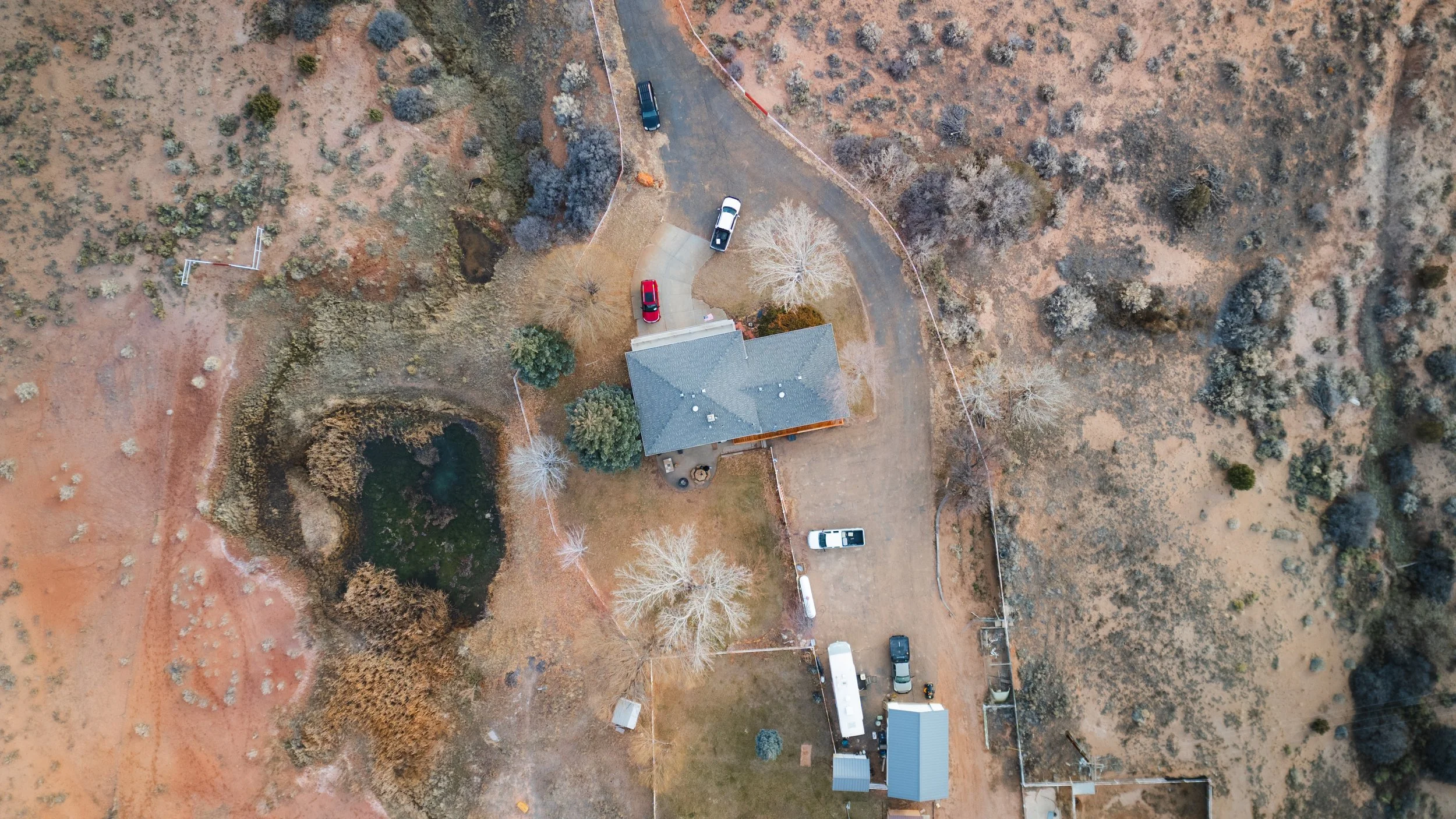 Aerial view of a house with a gray roof surrounded by trees, a pond, and a driveway with several parked vehicles in a rural area. Real estate drone photography and videography services in Roosevelt, Vernal, Uintah Basin, Utah, and nationwide.
