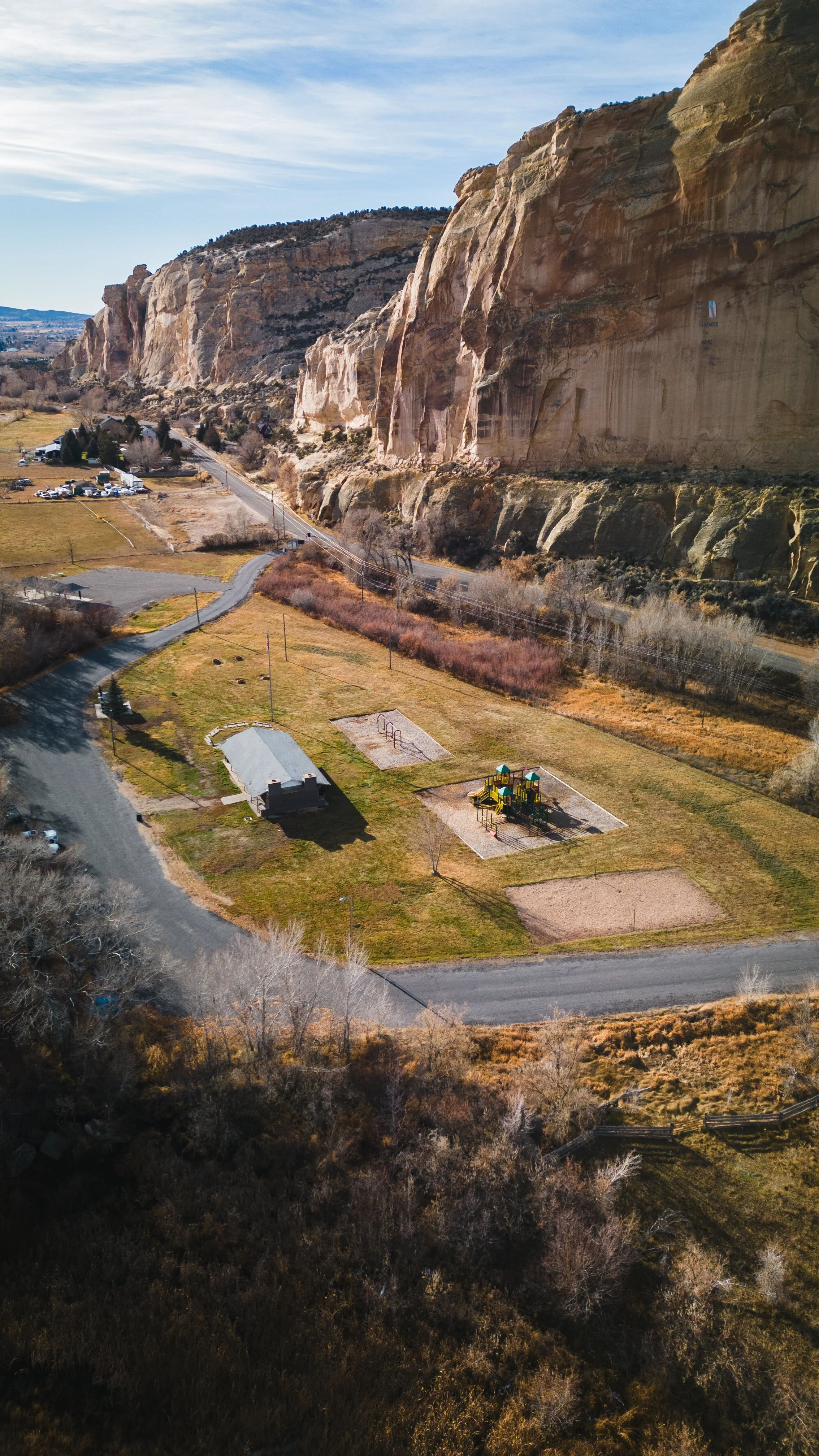 Aerial drone photograph view of a small park with playground, sand courts, and a pavilion, located at the base of large rock cliffs on a clear day. Drone photography and videography services in Roosevelt, Vernal, Uintah Basin, Utah, and nationwide.
