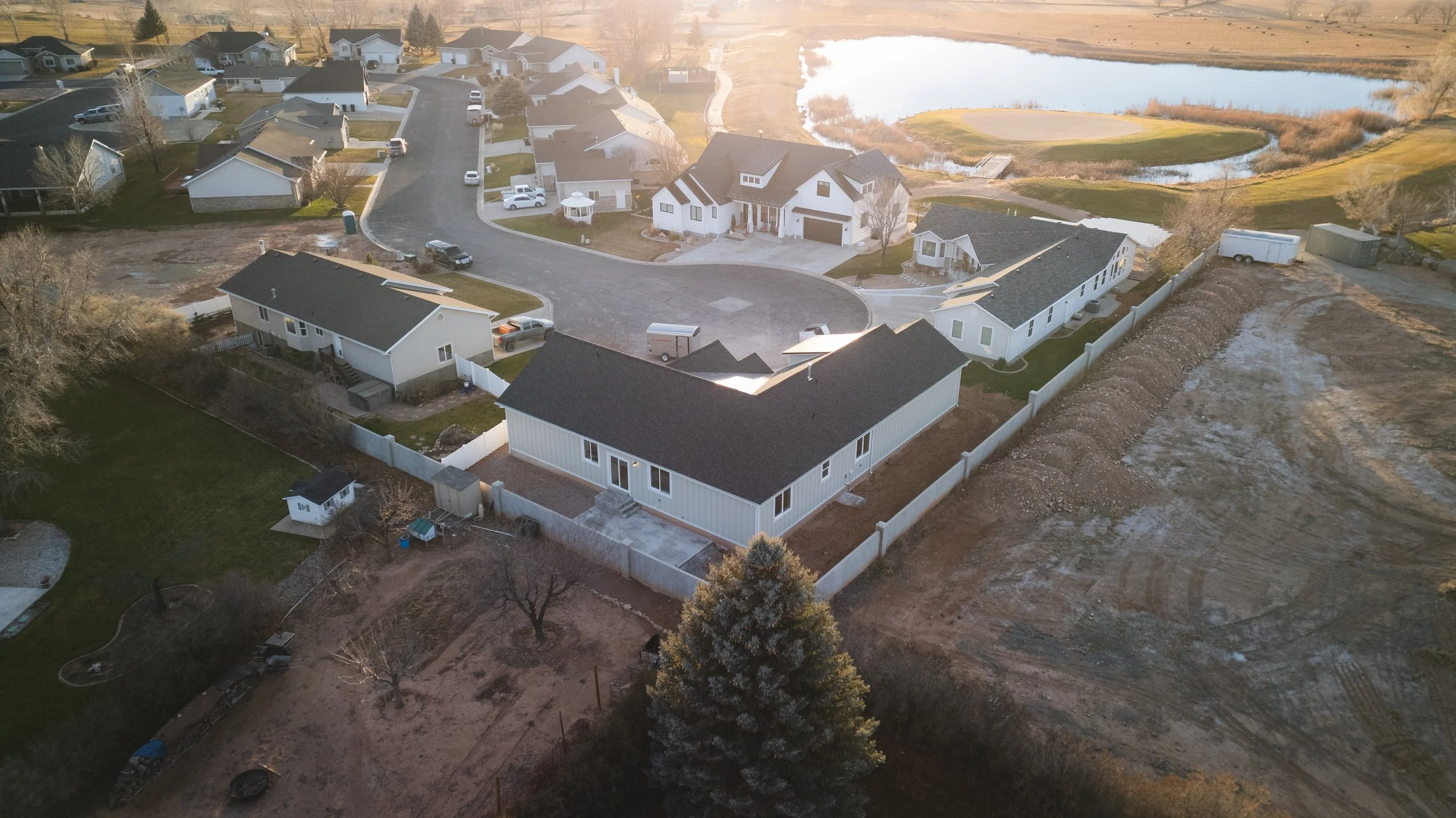 Aerial view of a neighborhood with several houses, a pond, and a golf course in the background, during late afternoon sunlight. Real estate drone photography and videography services in Roosevelt, Vernal, Uintah Basin, Utah, and nationwide,