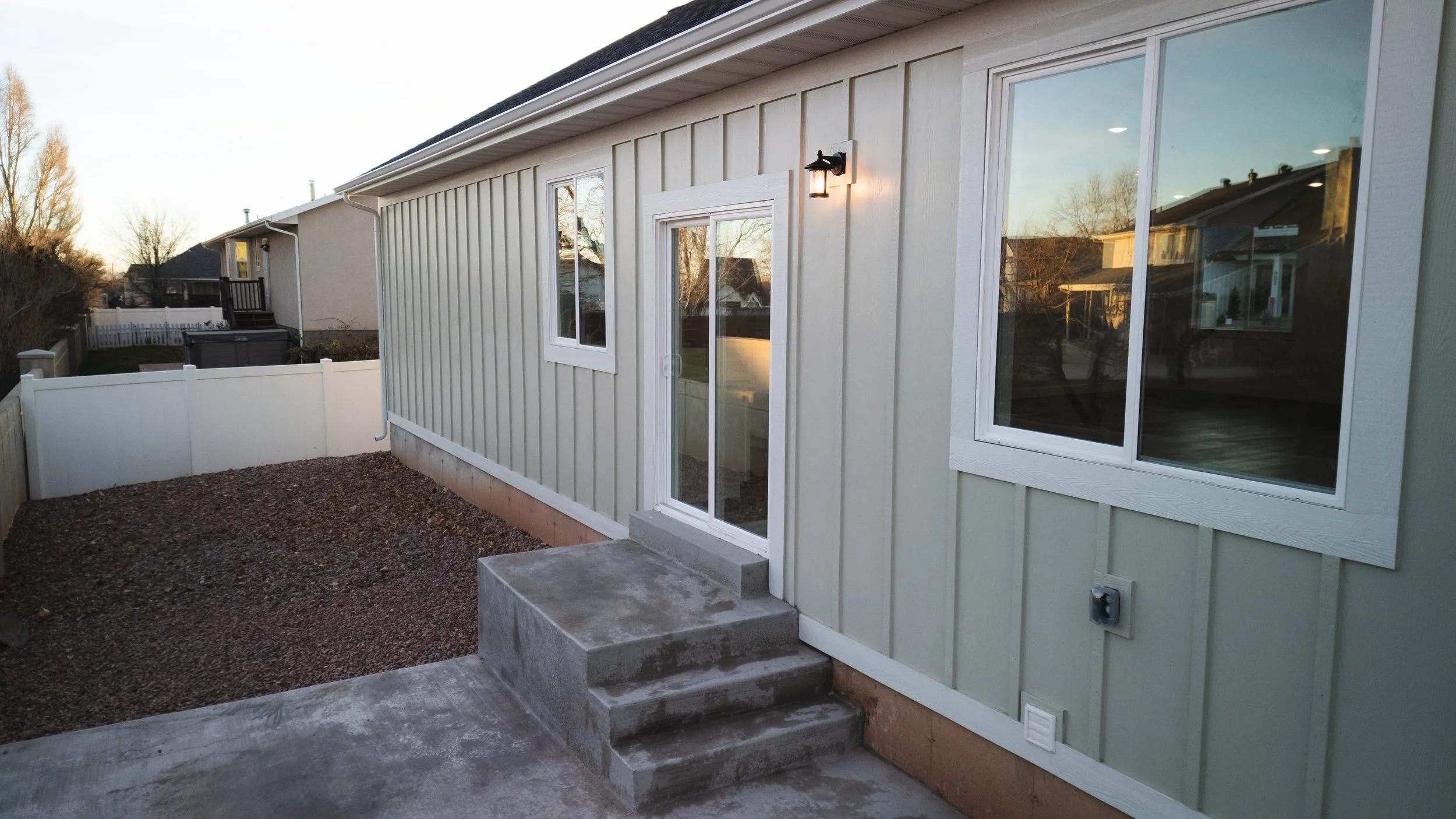 Backyard of a house with a small concrete step leading to a glass sliding door, beige siding, a wall-mounted light, and a fenced backyard. Real estate drone photography and videography services in Roosevelt, Vernal, Uintah Basin and nationwide.