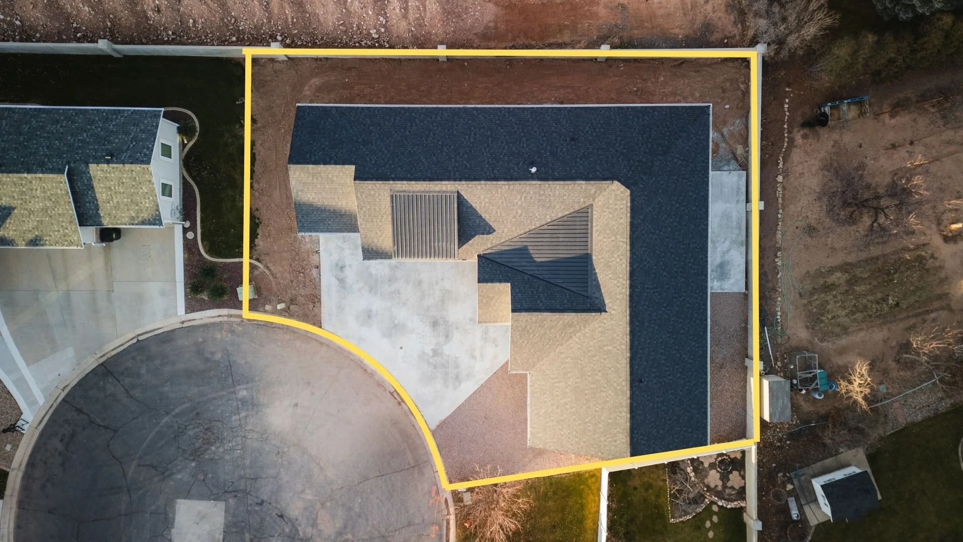 An aerial view of a house with a complex roof design, surrounded by a yard with a concrete patio and several trees, enclosed by a yellow boundary line. Real estate drone photography and videography services in Roosevelt, Vernal, Uintah Basin, Utah.