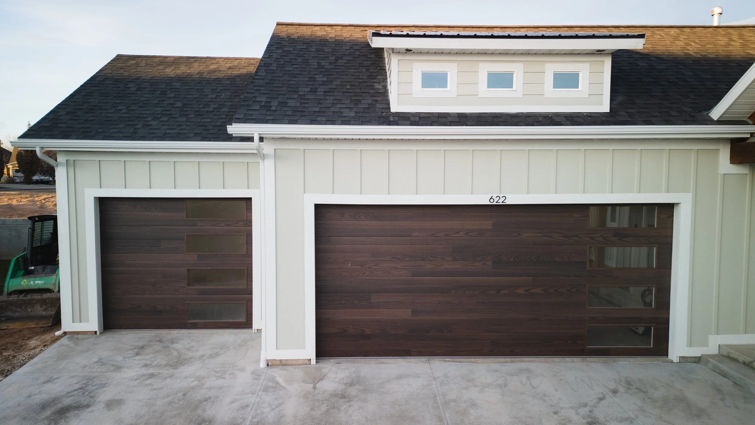Newly built house with two wood-paneled garage doors, black shingle roof, and small dormer window. Real estate drone photography and videography services in Roosevelt, Vernal, Uintah Basin, Utah, and nationwide.