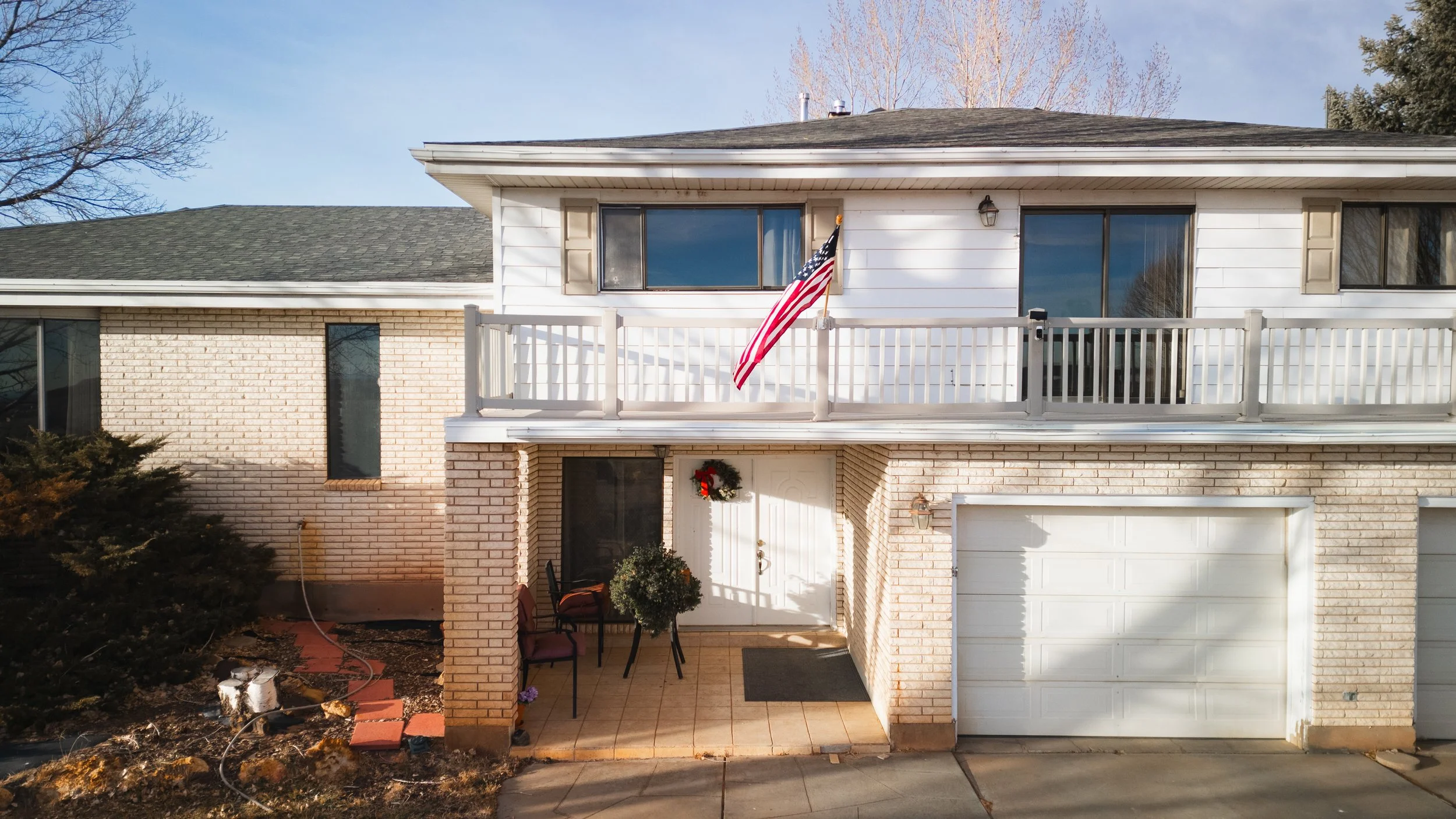 Front view of a two-story house with brick and white siding exterior, an American flag hanging from a balcony, a small porch, and a garage door. Real estate drone photography and videography services in Roosevelt, Vernral and Uintah Basin.