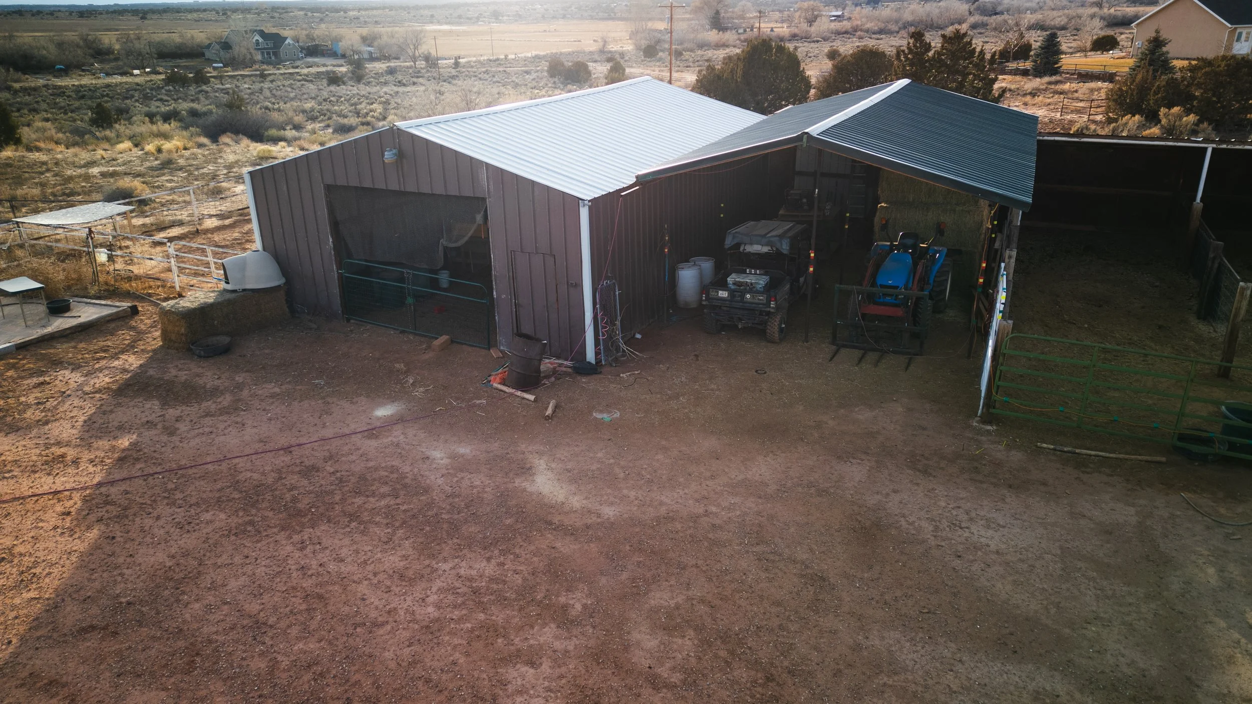 Aerial view of a farm shed with equipment, tractors, and a fenced yard in a dry, rural area with distant houses and open land. Real estate drone photography and videography services in Roosevelt, Vernal, Uintah Basin, Utah, and nationwide.