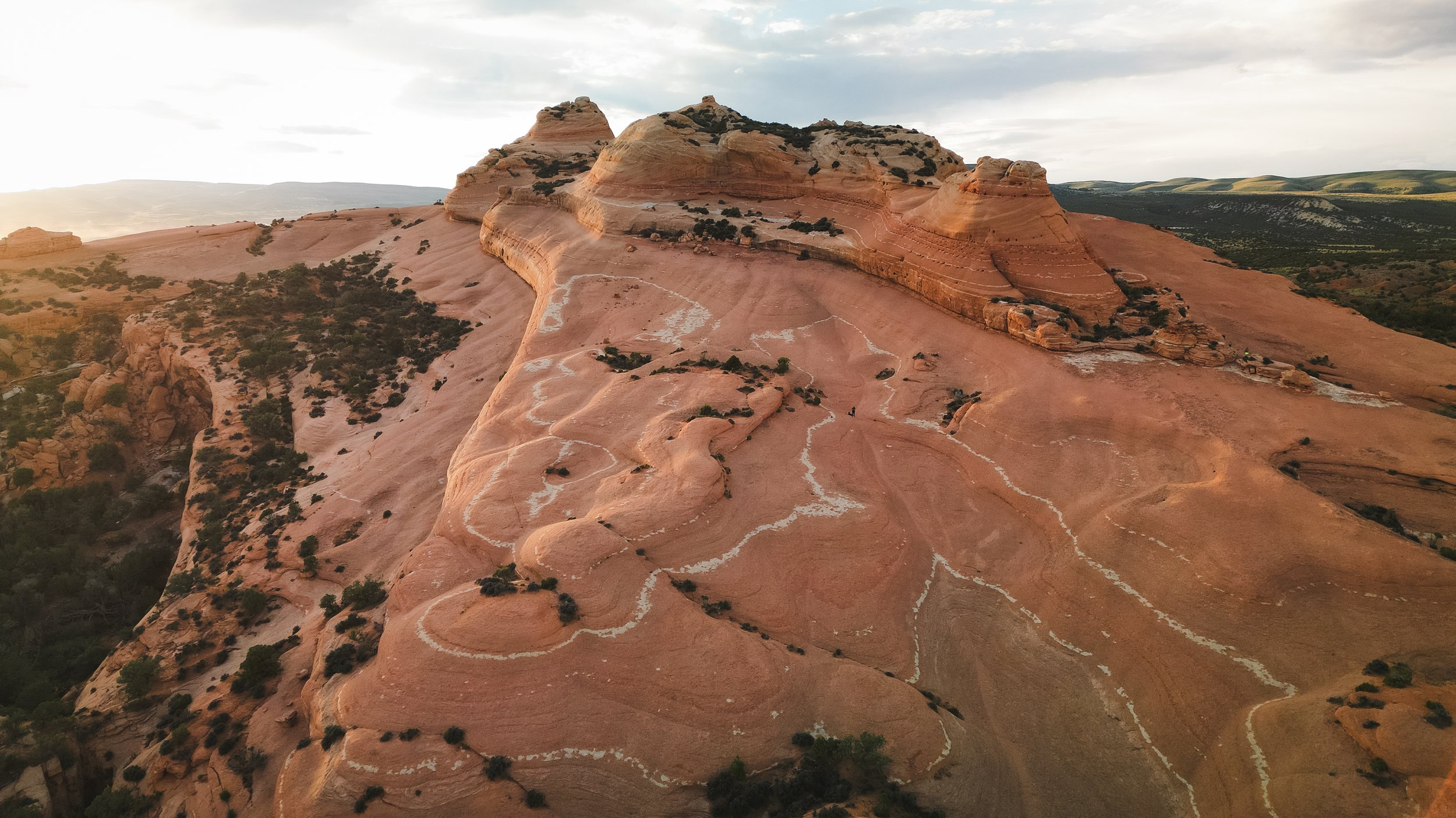 Aerial drone view of colorful rock formations and winding trails on a sandstone landscape, with sparse vegetation and a cloudy sky. Drone photography and videography services in Roosevelt, Vernal, Uintah Basin, Utah, and nationwide.