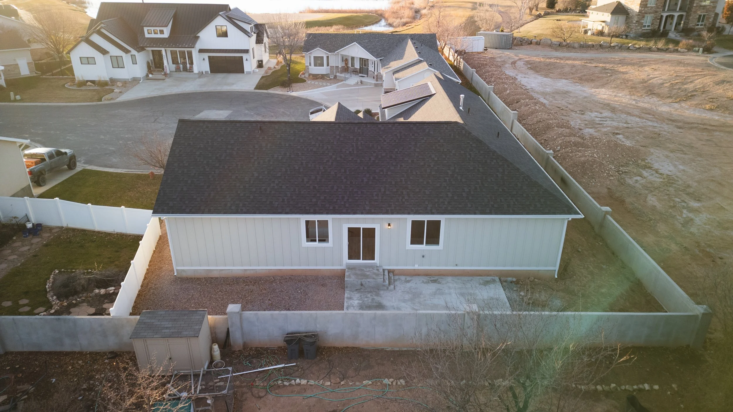 Aerial view of a new white house with gray roof, backyard with patio, and surrounding fences, neighboring houses, and dirt lot. Real estate drone photography and videography services in Roosevelt, Vernal, Uintah Basin, Utah, and nationwide.