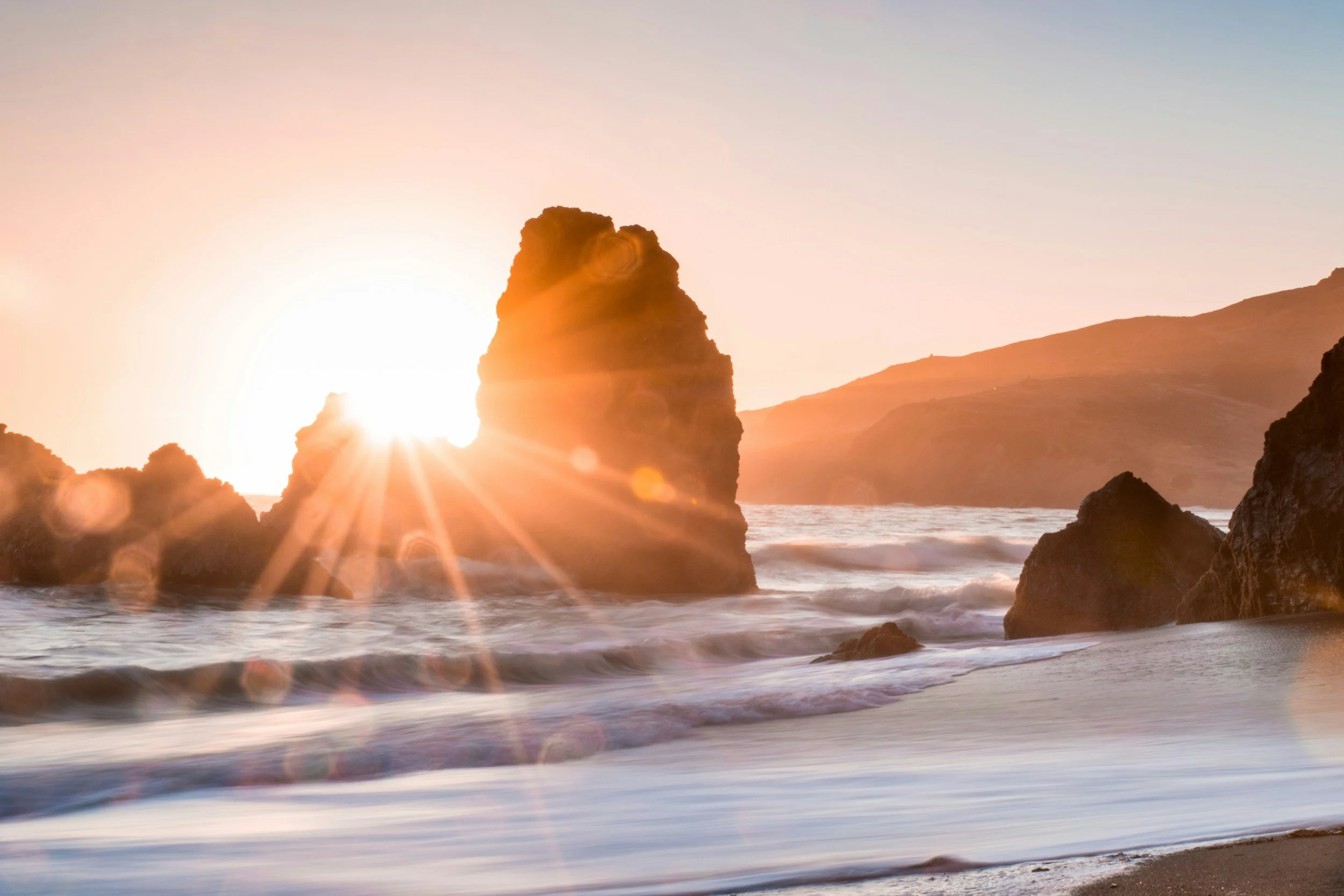 Sunset over rocky shoreline with waves crashing and a large rock formation silhouetted against the setting sun.