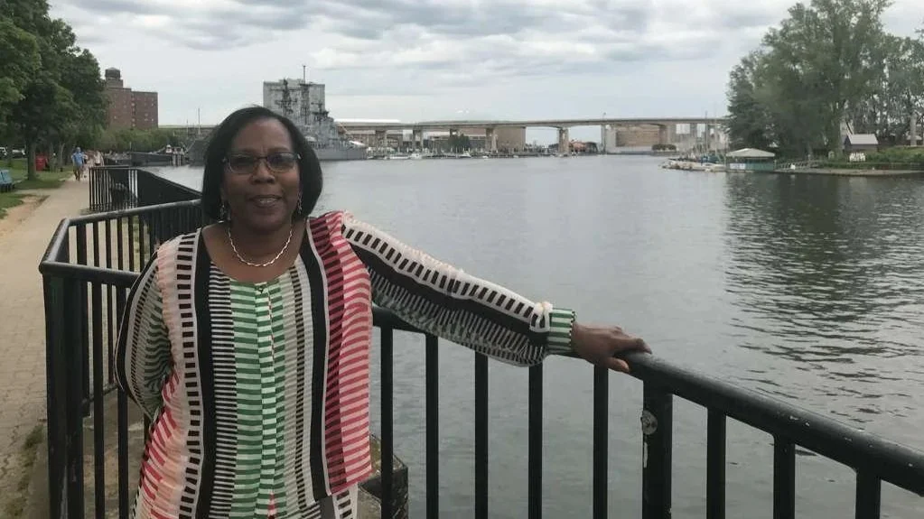 A woman standing by a waterfront railing with a river, trees, and a bridge in the background on an overcast day.