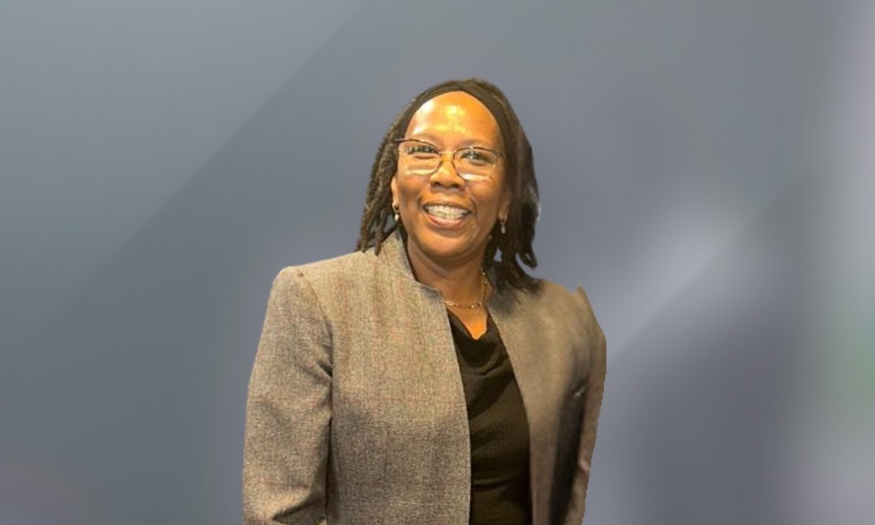 A woman with glasses and shoulder-length dreadlocks, wearing a gray blazer over a black top, smiling in front of a plain gray background.