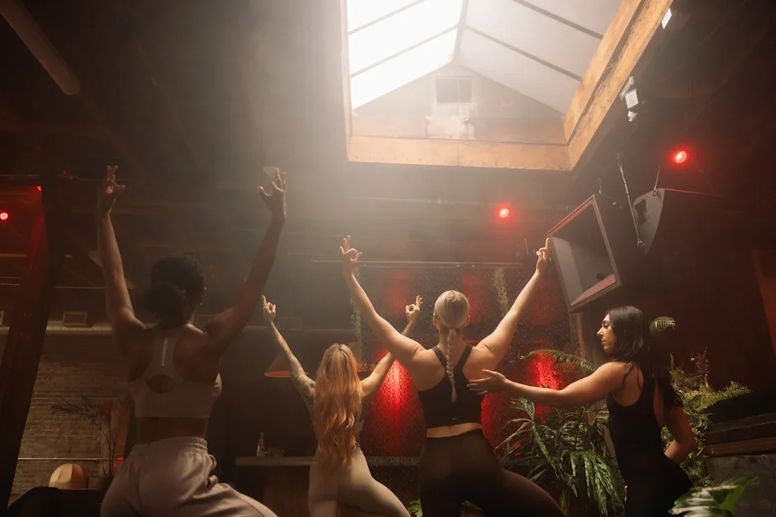Four women dancing with their arms raised in a dimly lit indoor space with a skylight overhead.