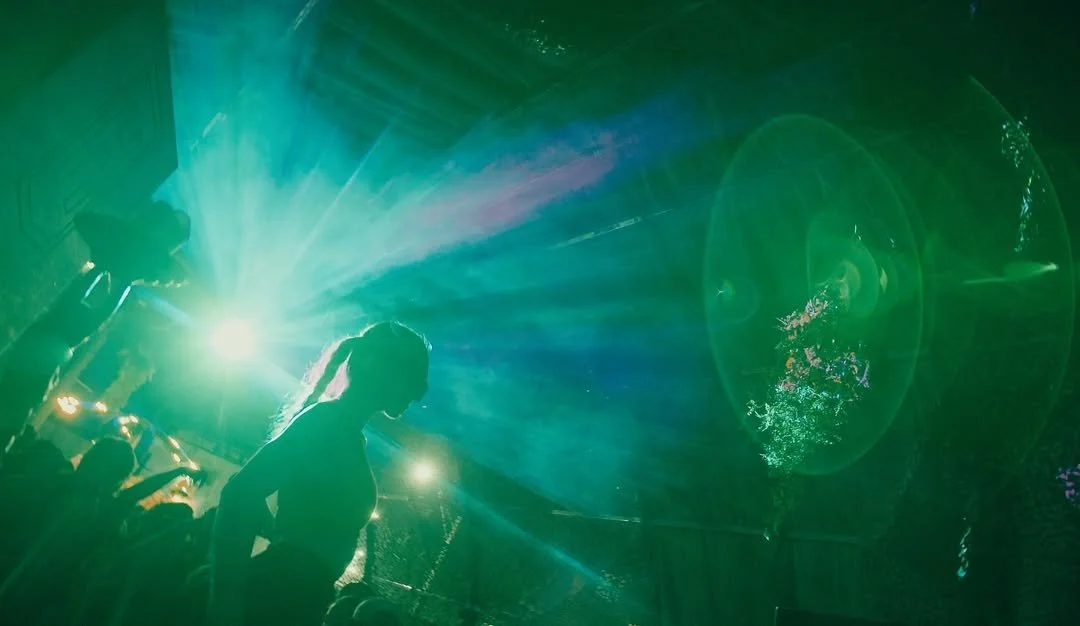Silhouette of a person with colorful laser lights and water fountains in the background, likely at a nightclub or outdoor event.