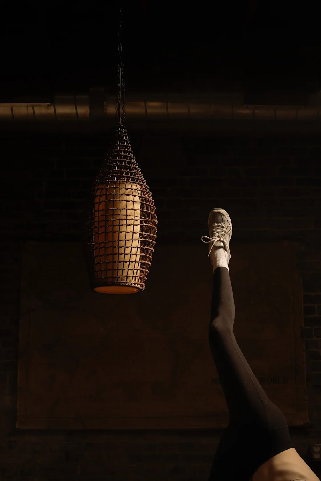 A person raising one leg, wearing a white sneaker and black leggings, in front of a dark background with a hanging decorative light fixture.