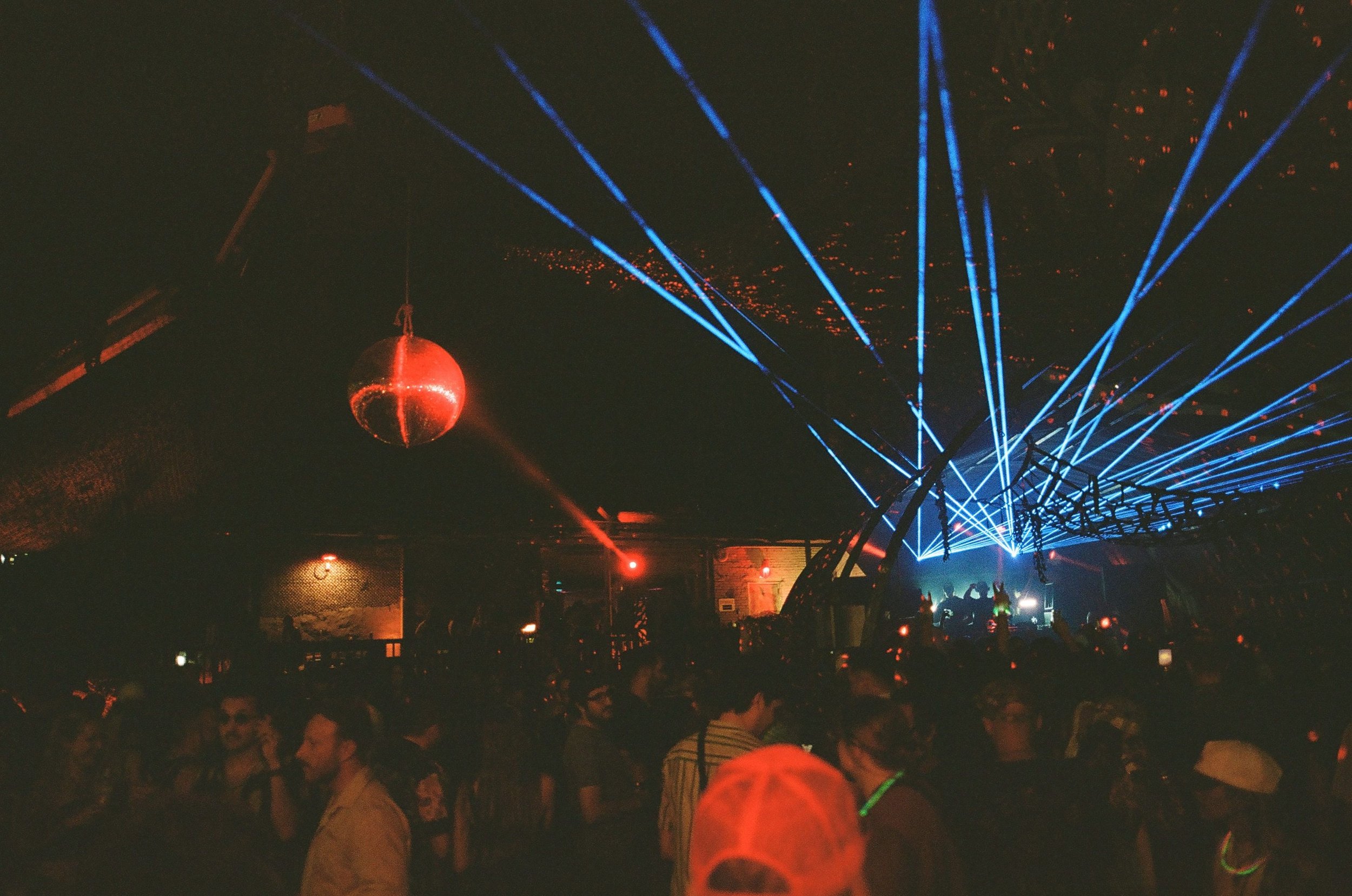 Nightclub scene with a crowd of people, blue laser lights, a red disco ball, and a DJ on stage.