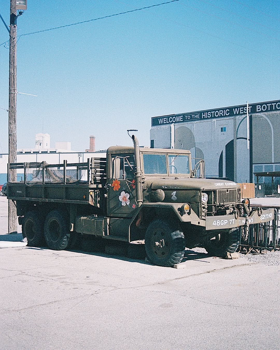 A vintage military-style truck painted in camouflage with flower decals on the door, parked in front of a building with a sign that reads 'Welcome to the Historic West Bottoms'.