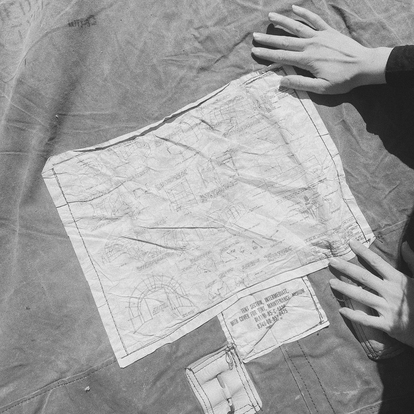 A person with light skin is examining a large, crumpled blueprint or technical drawing spread out on a table or surface. The blueprint contains various architectural sketches and diagrams, including a tent or dome structure, with some text and measur