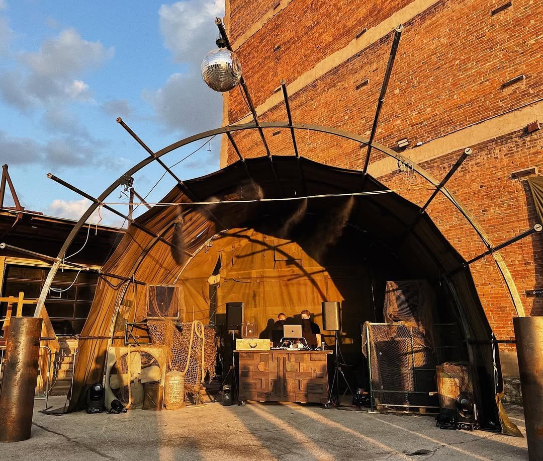 An outdoor stage set up against a brick wall with a DJ booth and speakers, decorated with gold and animal print fabrics, and a disco ball hanging above.