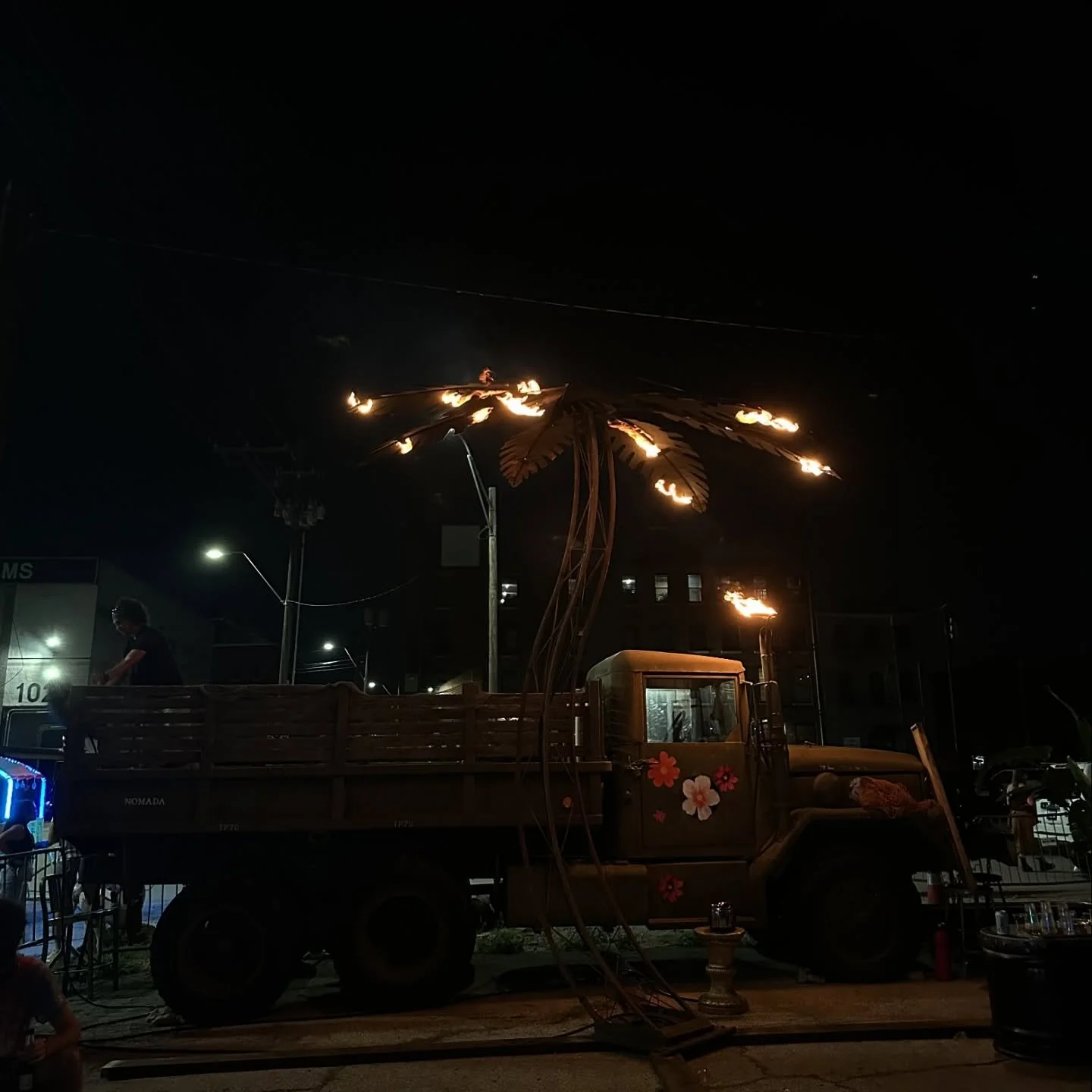 A decorated truck with large flaming palm tree sculpture on top, illuminated against a dark night sky, in an outdoor setting with people and streetlights.