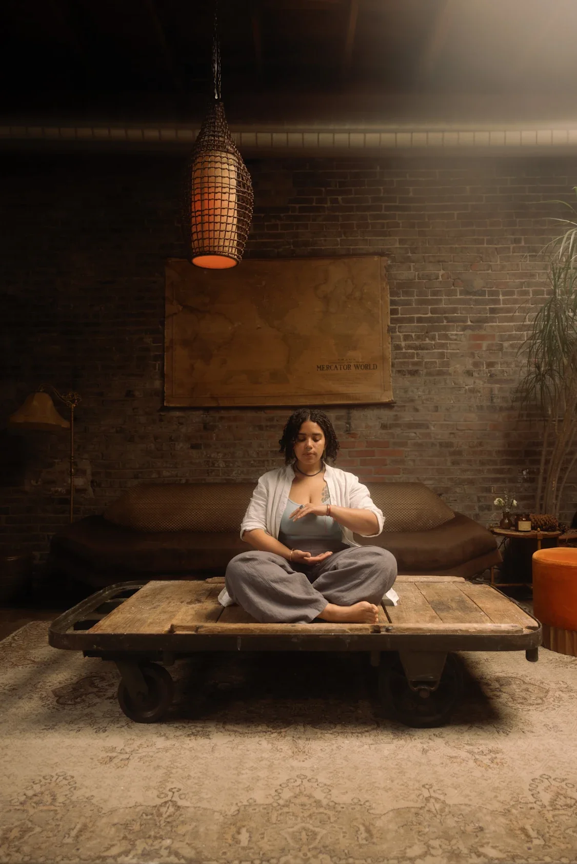 A woman sitting cross-legged on a wooden cart in a cozy, dimly lit room with brick walls. She is practicing meditation or mindfulness, with her eyes closed and one hand on her wrist. There's a sofa behind her, a large world map on the wall, and vario