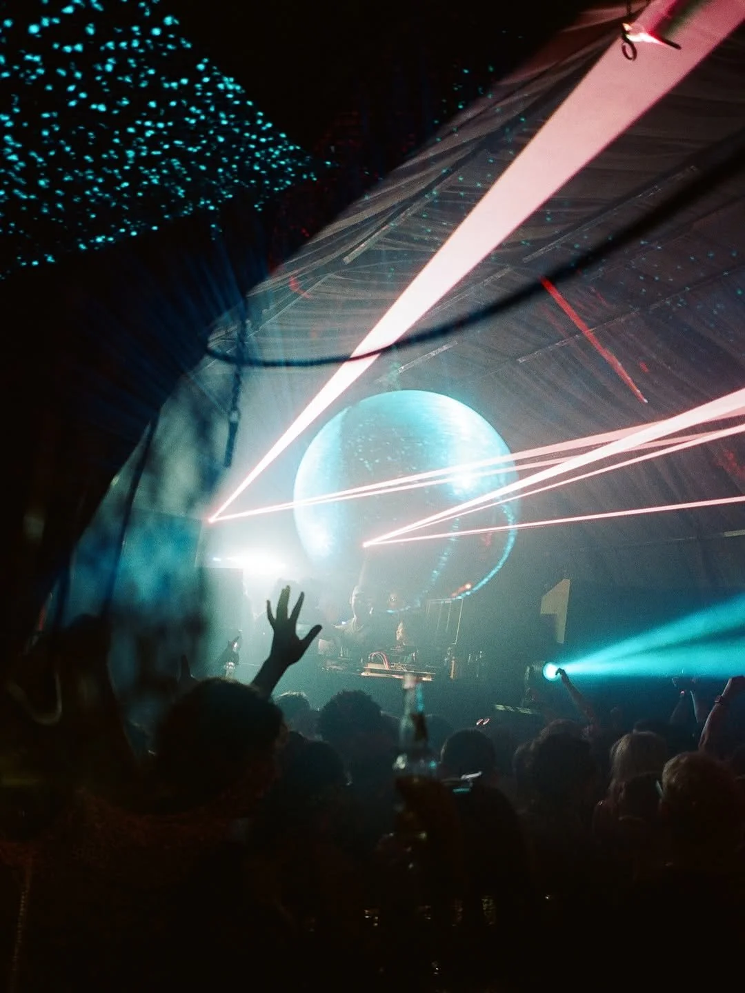 A concert or nightclub scene with a large illuminated globe on stage, laser lights, and a crowd with raised hands and drinks.