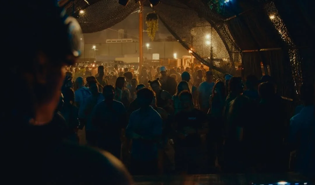 Crowd of people gathered at a night event under a large tent with dim lighting and some hanging decorations.