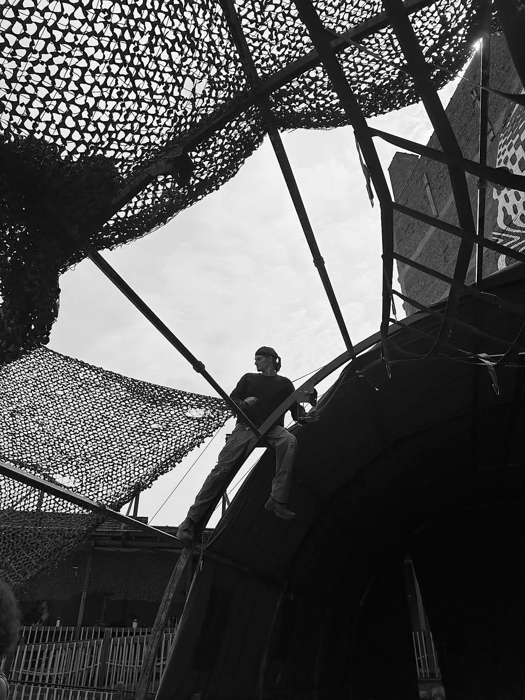 A person sitting on the edge of a metal structure with camouflage netting above, looking to the side, with a cloudy sky in the background.
