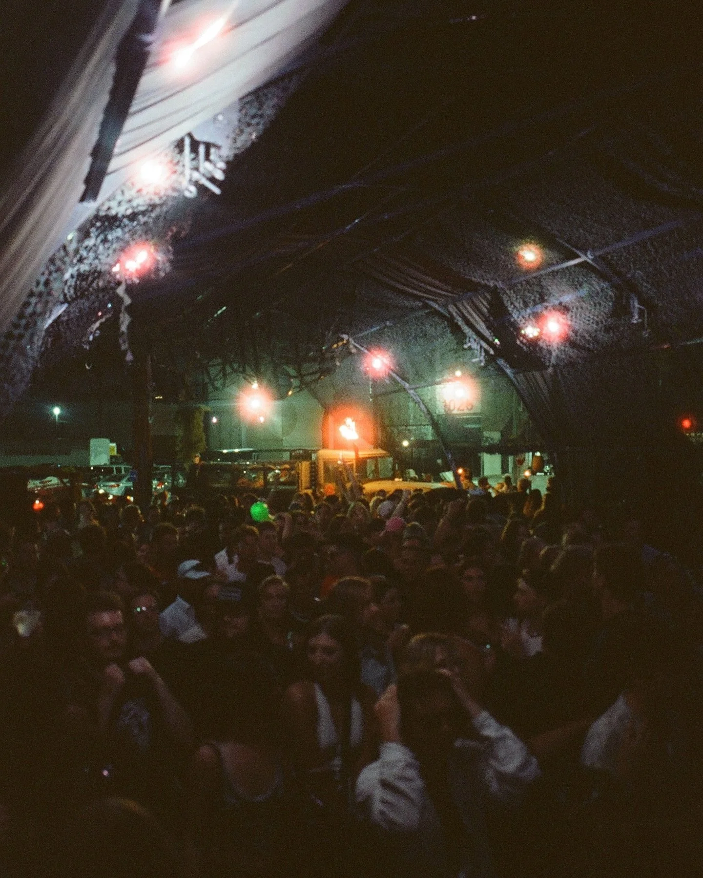 Nighttime outdoor concert scene with a large crowd of people seated and standing, illuminated by stage and ambient lights, with a dark sky and some structures visible in the background.