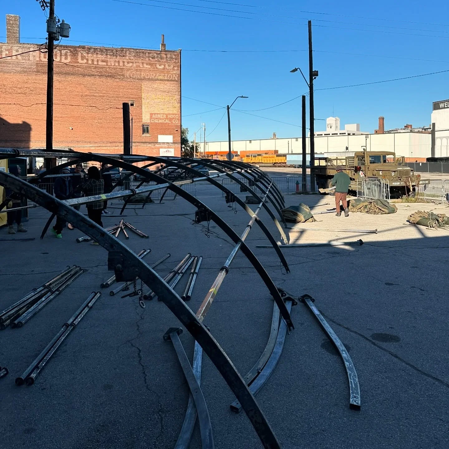 People assembling or disassembling stage structures outdoors on a paved lot, with metal frames and poles scattered around, and some equipment and a military truck nearby. Buildings and train cars are visible in the background under a clear blue sky.